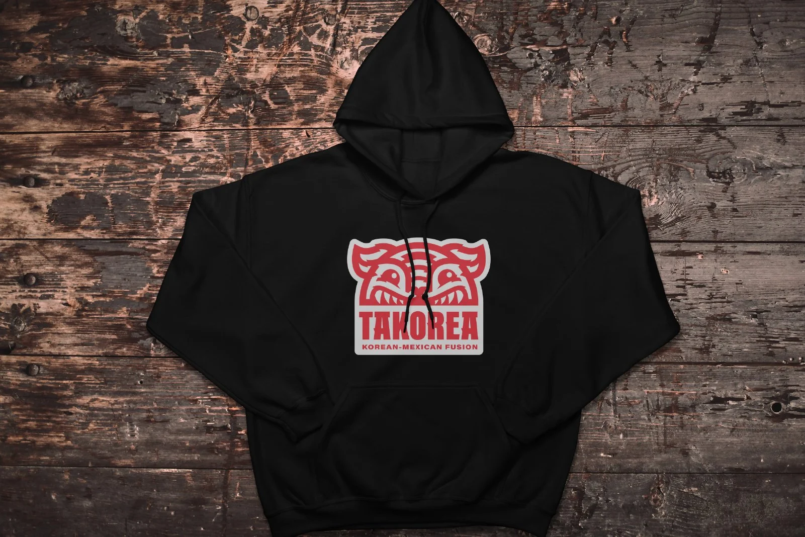 Black hoodie with a red and white logo that resembles a stylized dragon or creature, with the text "TAKOREA" and "KOREAN-MEXICAN FUSION" on the front, laid on a rustic wooden surface.