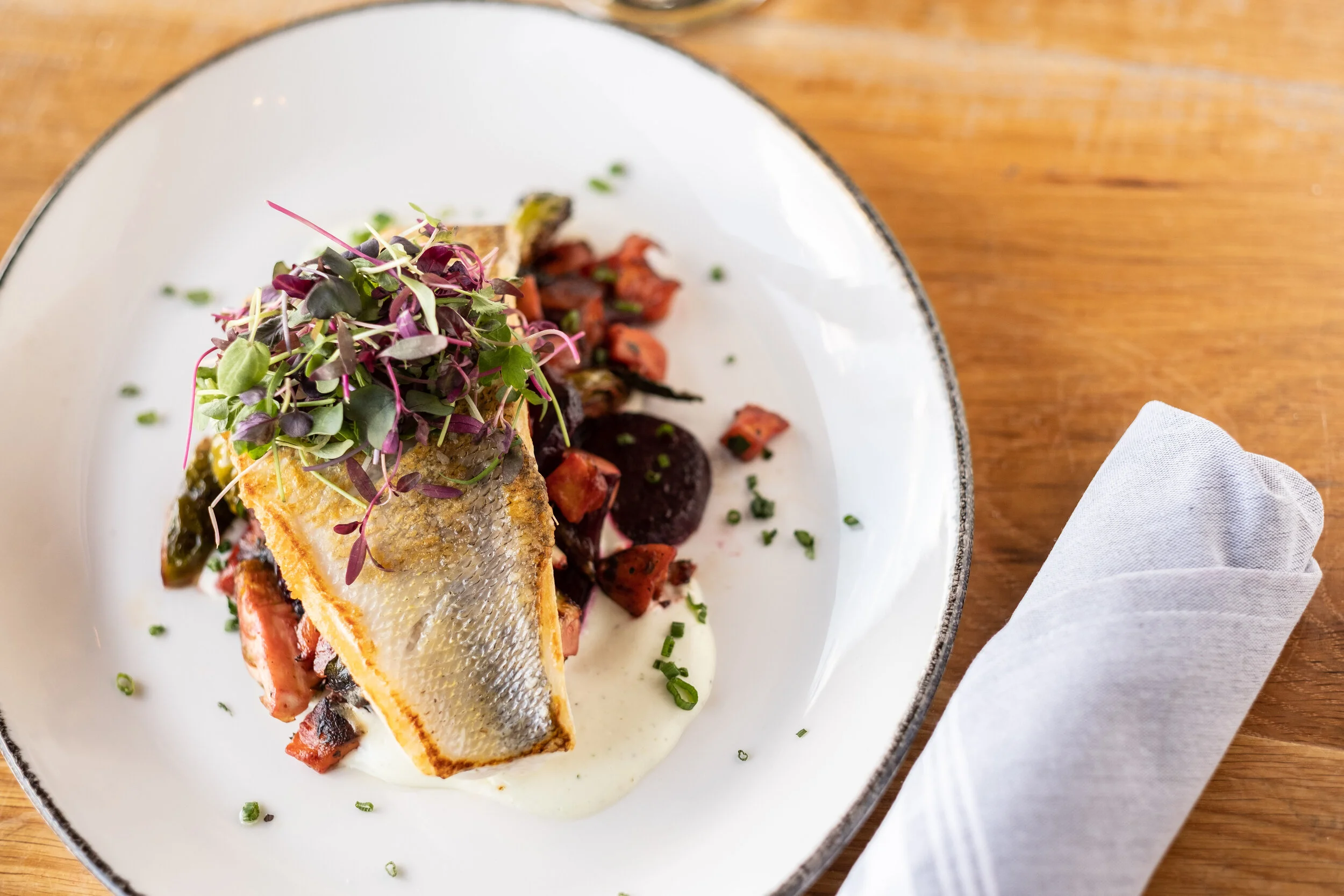 A plated dish featuring a grilled fish filet on a bed of diced tomatoes, beets, and greens, garnished with microgreens, served with a creamy white sauce, on a white plate with a black rim, placed on a wooden table with a white napkin.