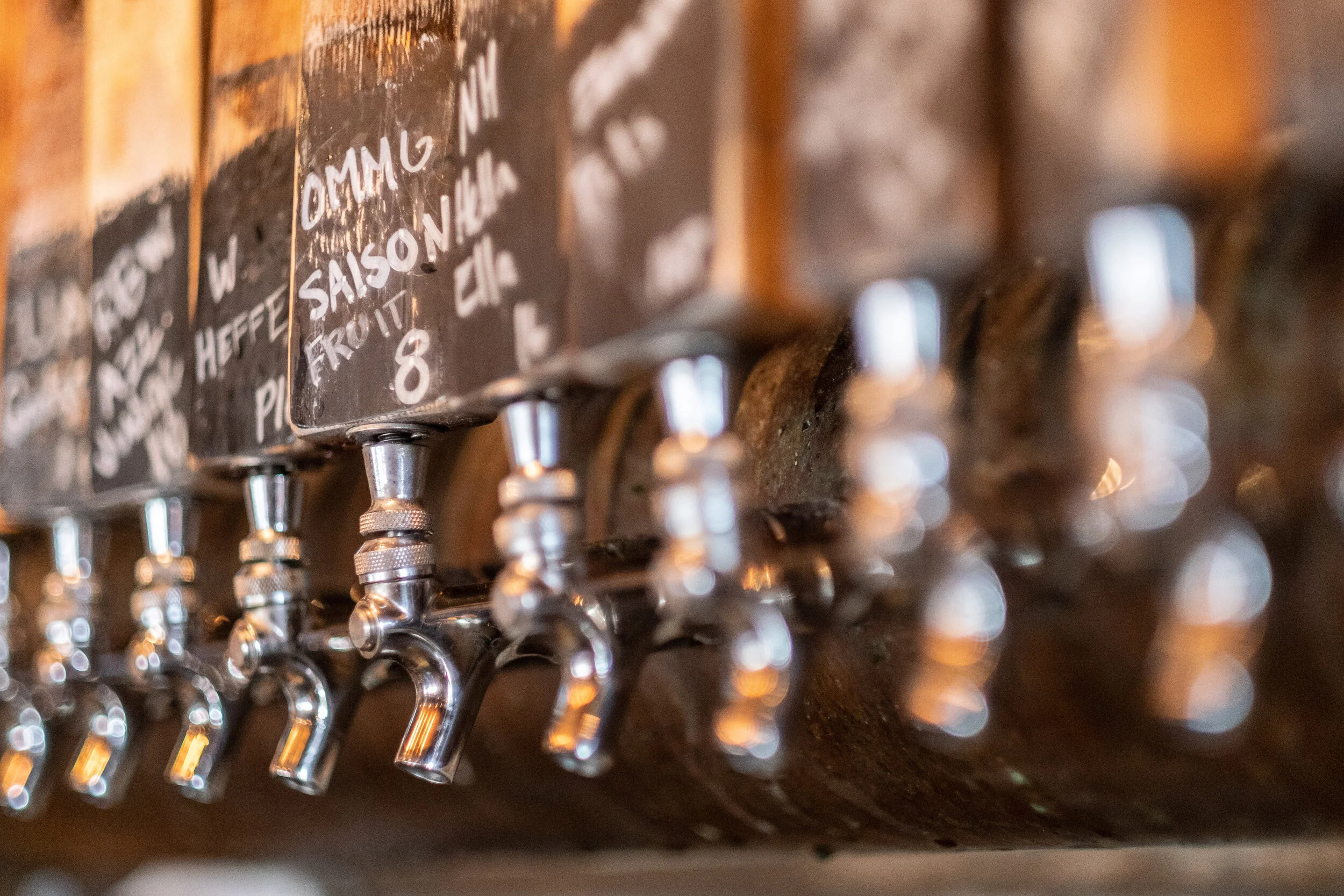 Close-up of brass beer taps attached to a wooden wall at a bar, with a chalkboard menu above listing various beers and their prices.