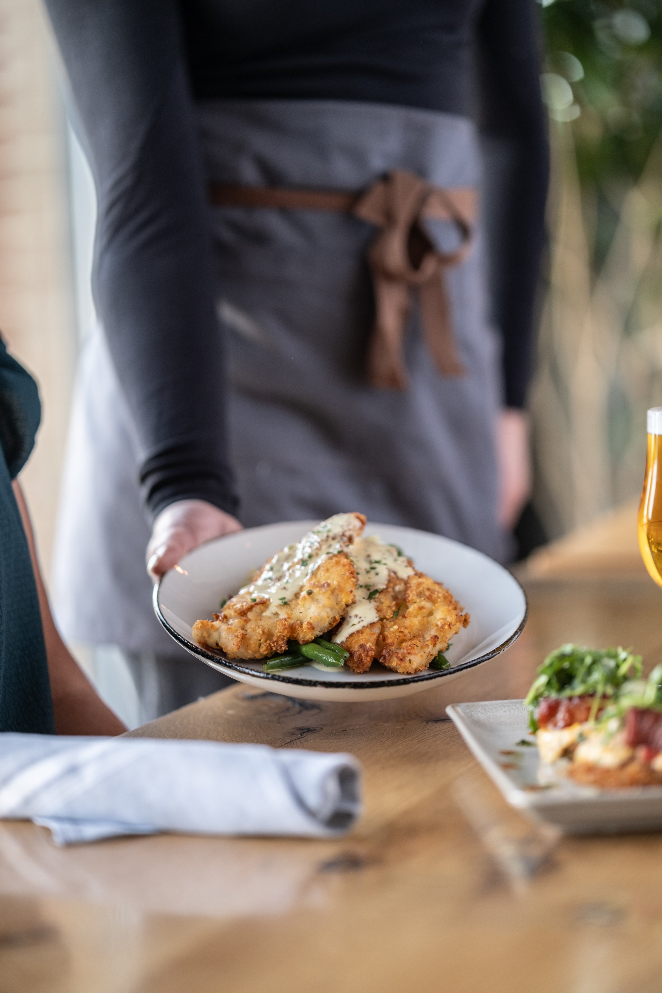 Waiter serving fried fish fillets with white sauce and green beans on a plate at a restaurant table.