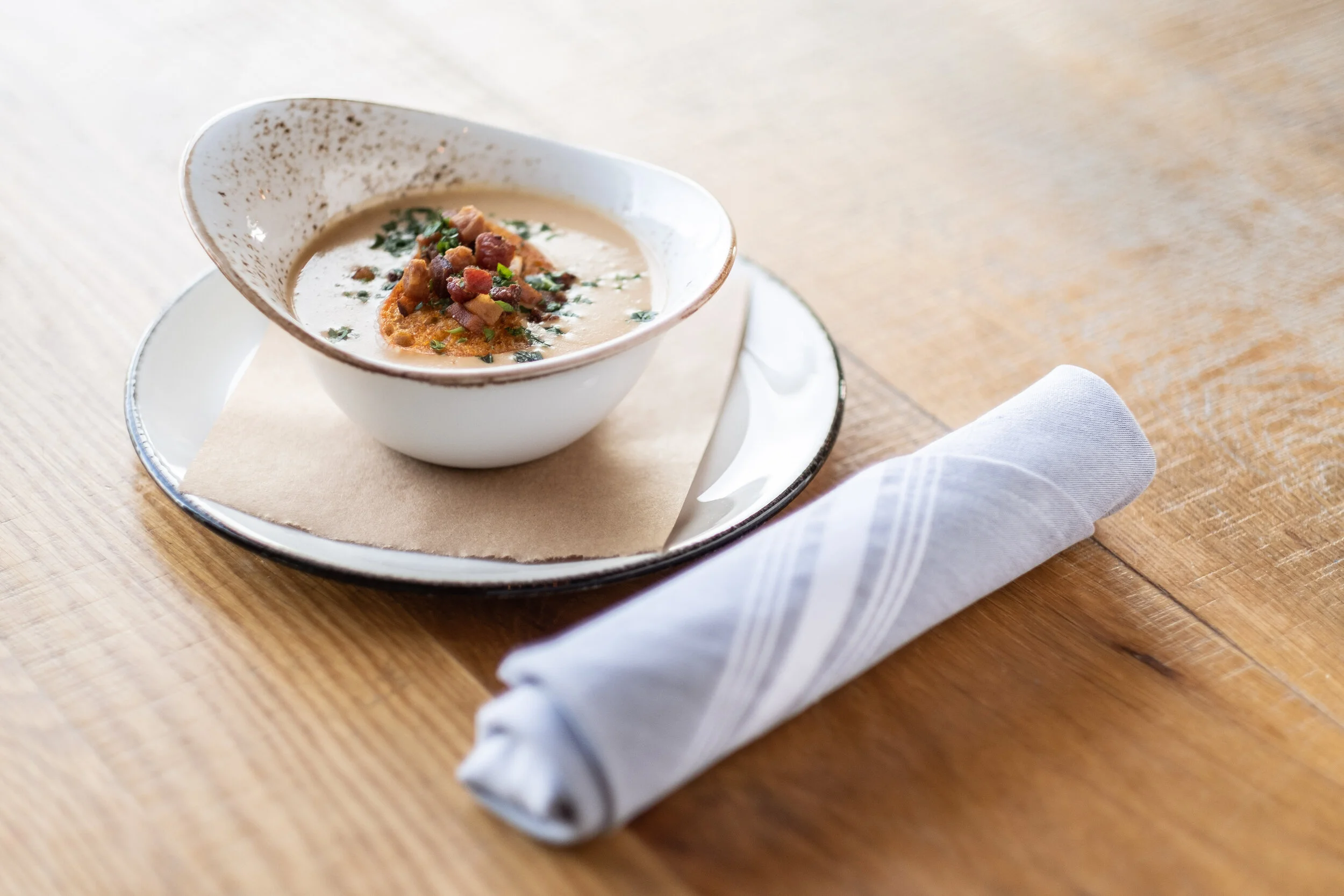 A bowl of creamy soup garnished with chopped herbs and bacon bits on a wooden table next to a rolled white napkin.