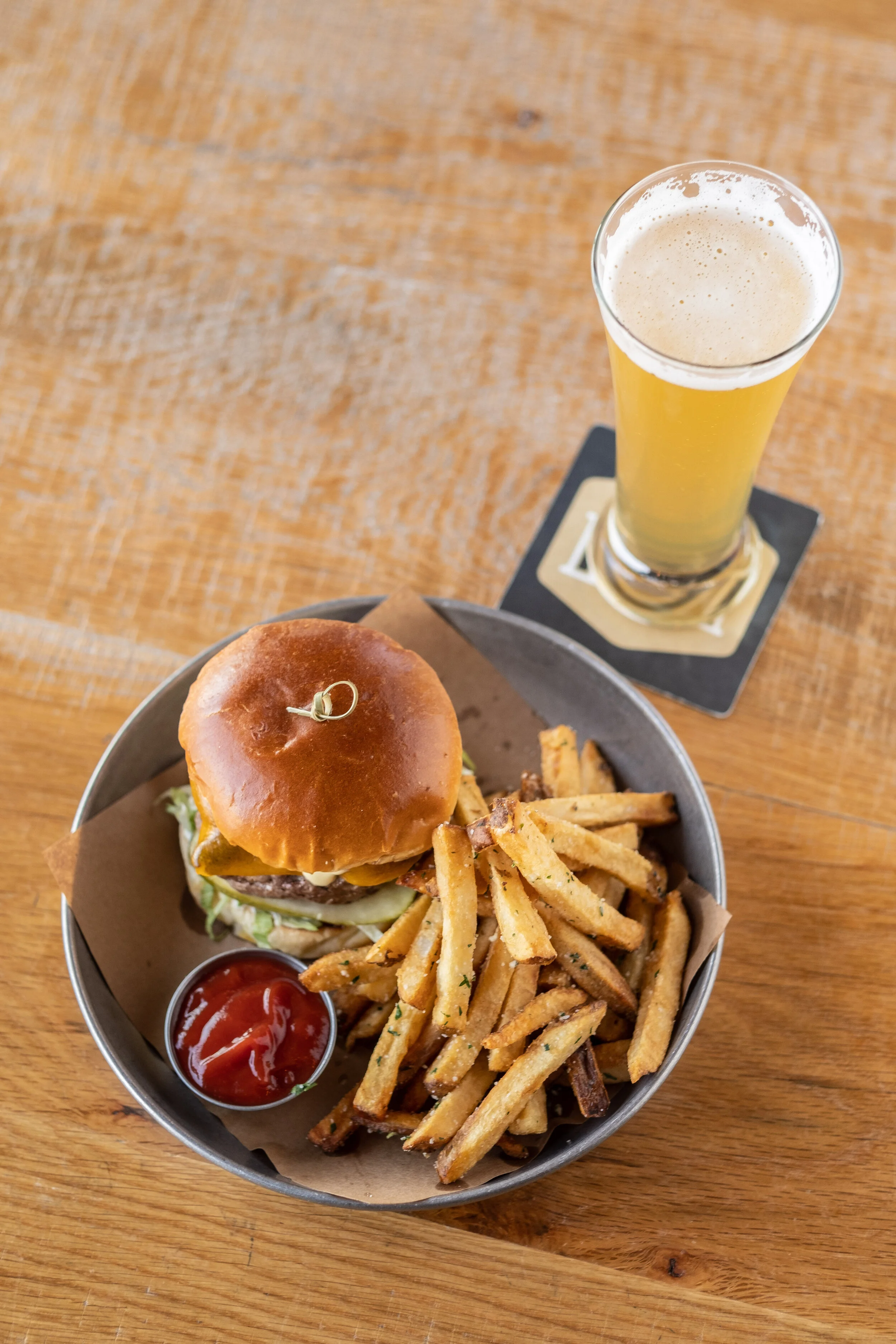 A burger with fries, ketchup, and a glass of beer on a wooden table.