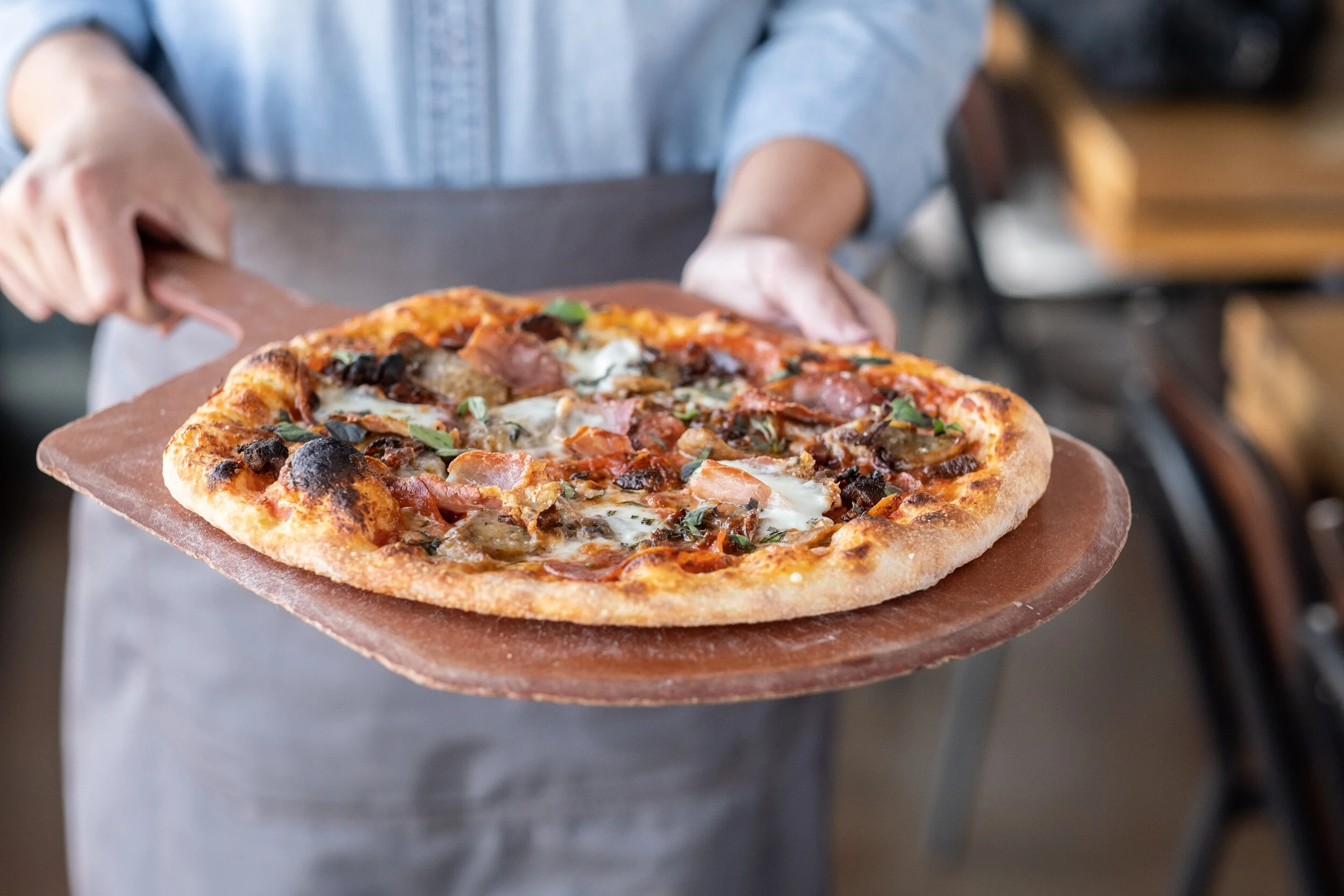 Person holding a wooden pizza peel with a freshly baked pizza topped with cheese, meats, and vegetables in a restaurant setting.