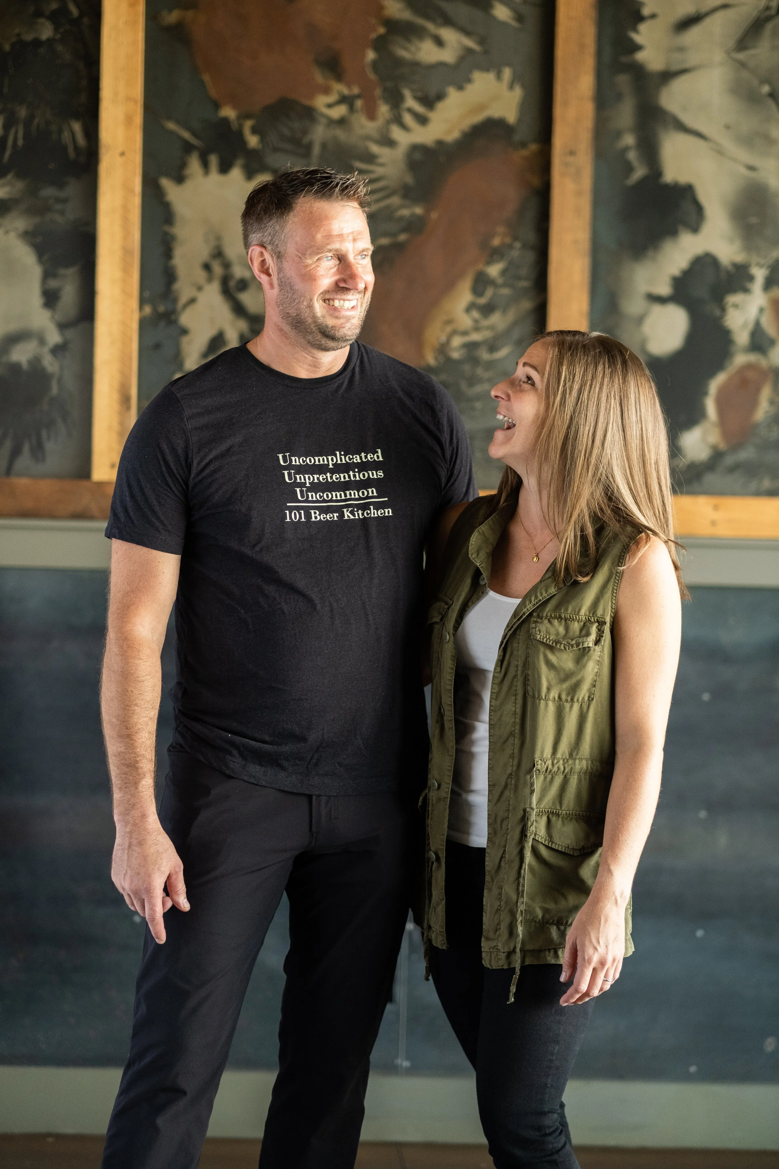 A man and woman standing close together inside a brewery or bar, smiling and looking at each other, with colorful abstract art on the wall in the background.