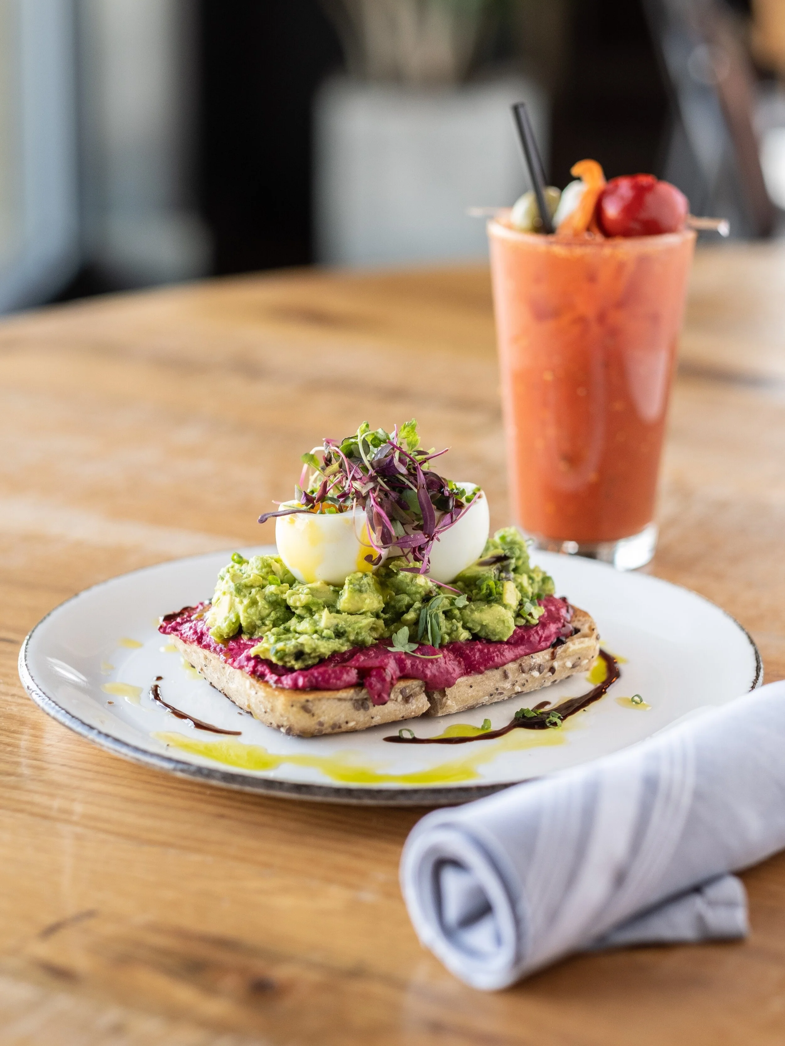 A plate with avocado toast topped with a halved boiled egg and microgreens, garnished with balsamic glaze and olive oil, and a tall glass of pink smoothie with fruit garnishes in the background on a wooden table.