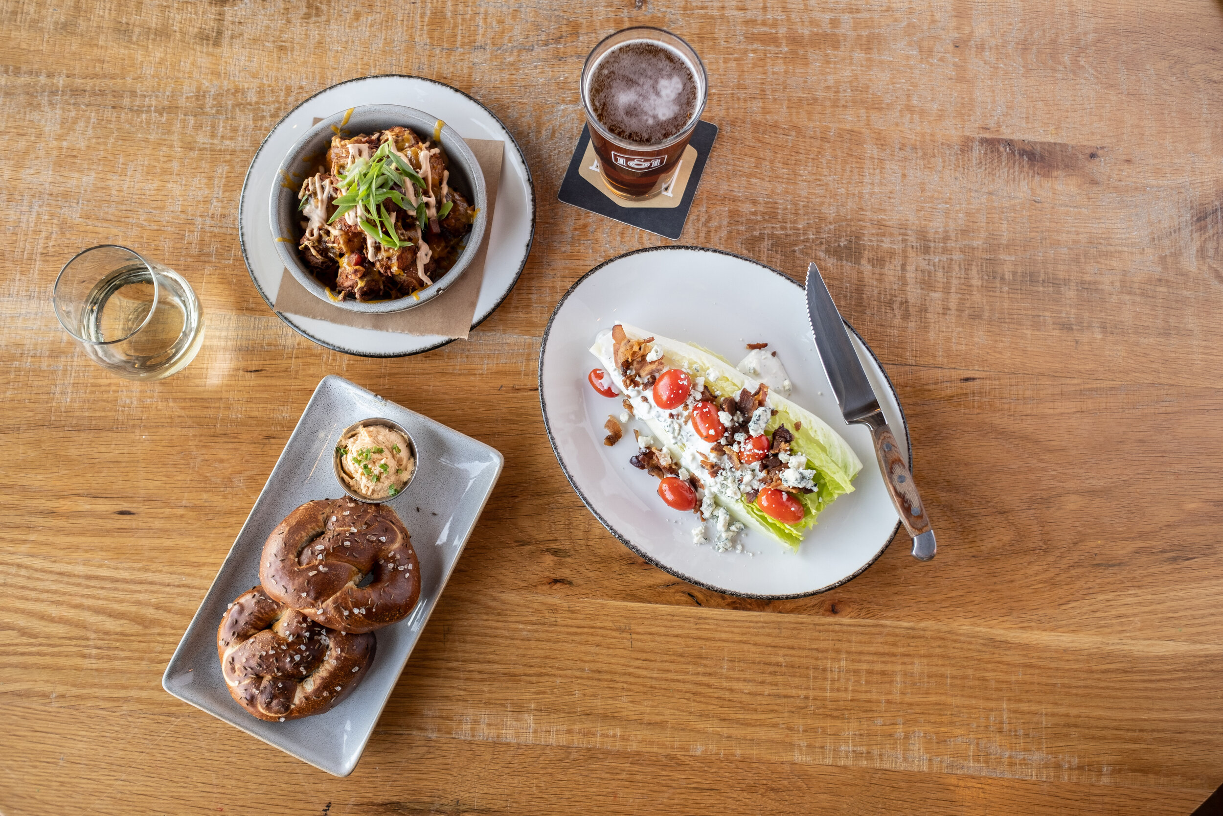A top-down view of a wooden table with various dishes including a salad with cherry tomatoes, a pretzel with mustard, a bowl of beef stew garnished with green onions, a glass of white wine, and a glass of dark beer.