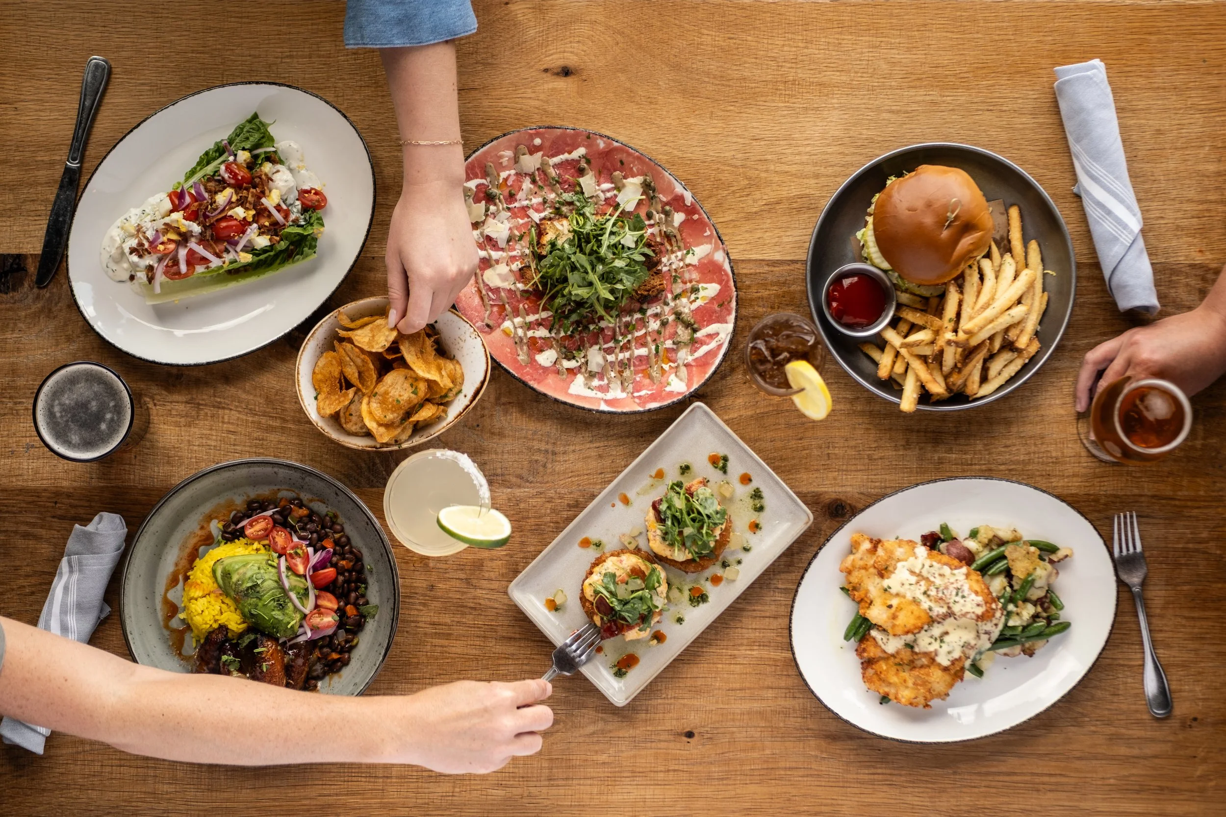 A top-down view of a wooden table with various dishes including a salad, potato chips, a carpaccio, fries with a burger, a plate of pulled pork, and a plate of chicken with vegetables, along with drinks and utensils.