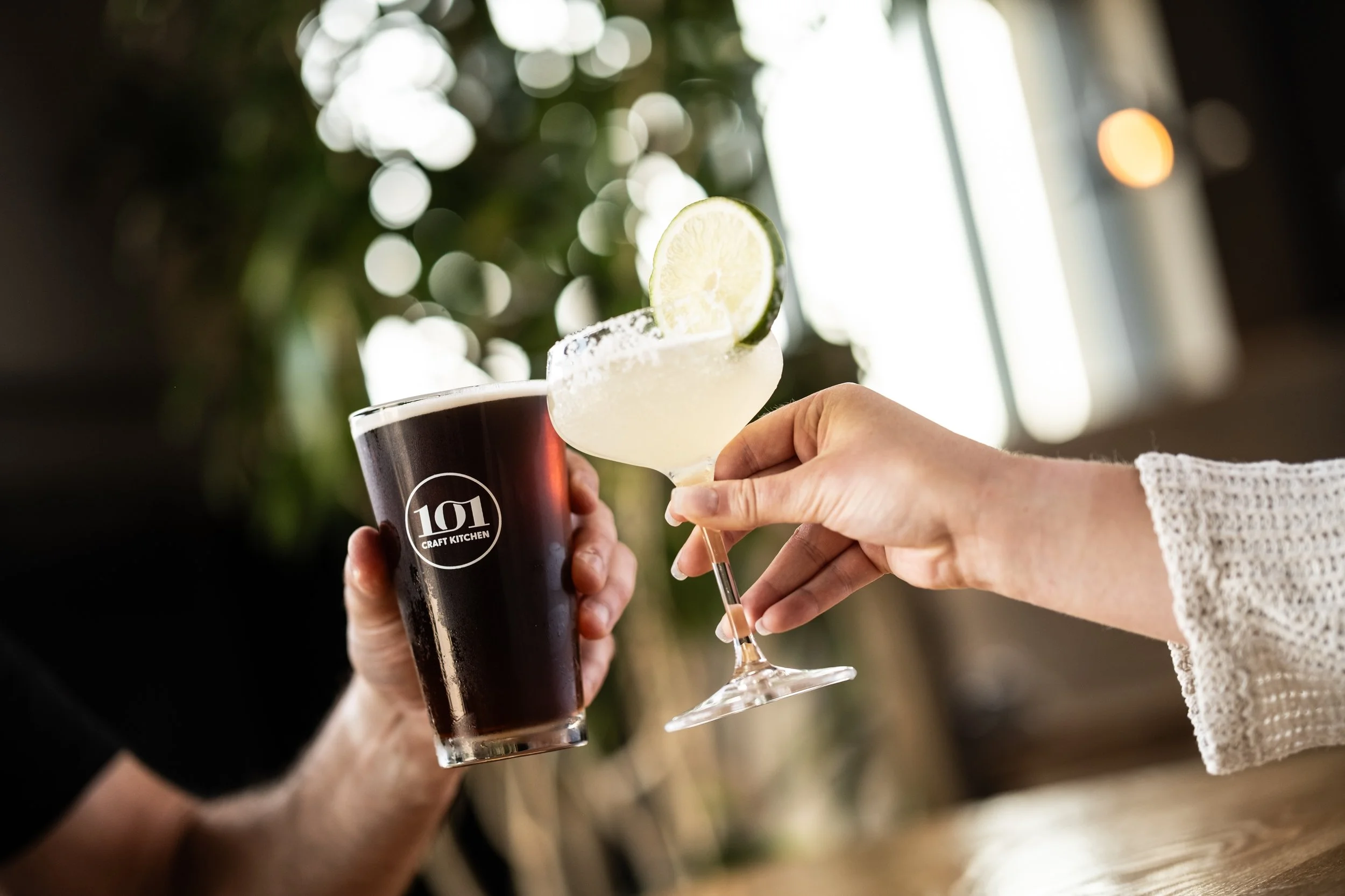 Person holding a cocktail glass with a lime wedge, salt rim, and a drink, next to a person holding a beer with a "101 Craft Kitchen" logo, in a cozy bar or restaurant setting.