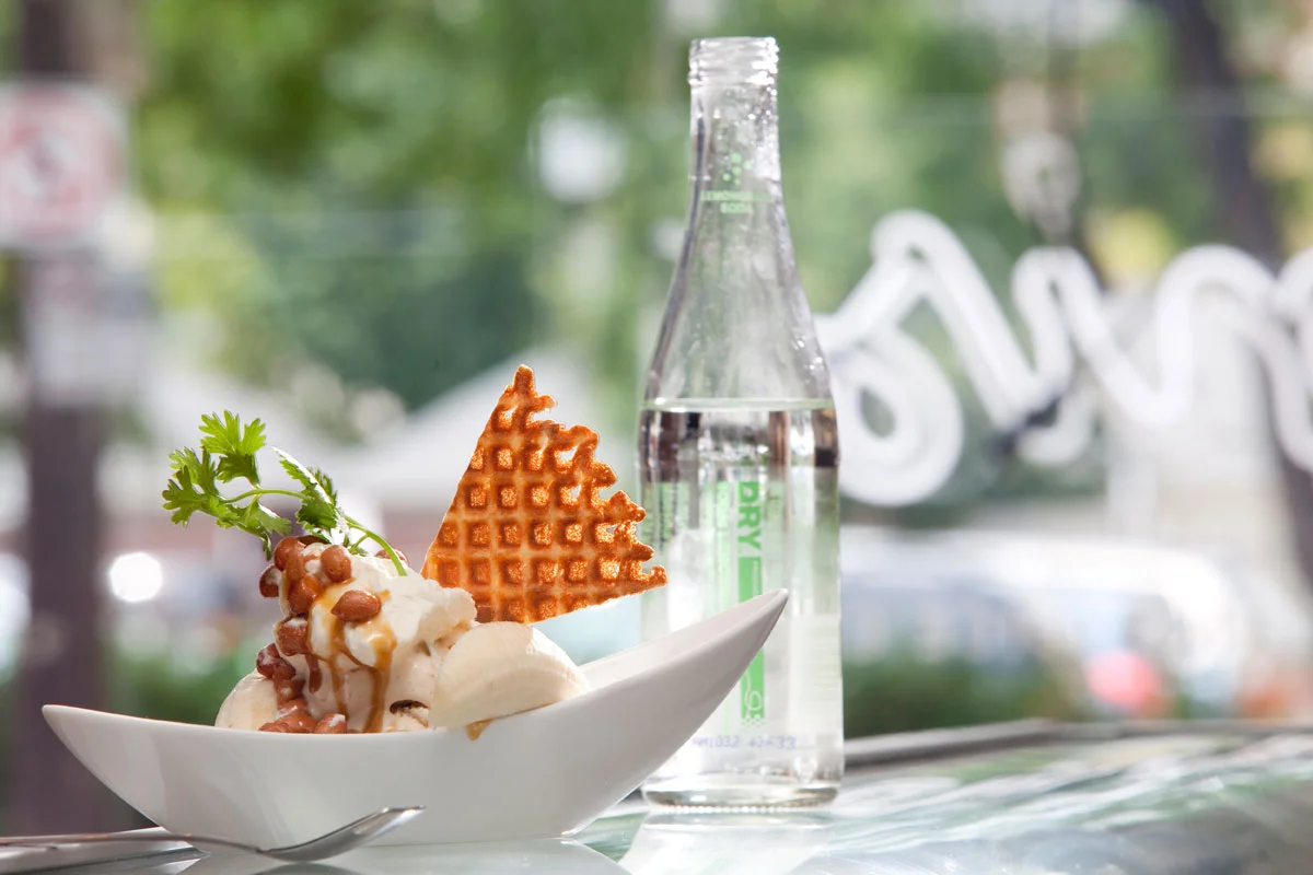 Ice cream topped with caramel sauce, chocolate sprinkles, a sprig of parsley, a waffle cone, and a bottle of sparkling water on a table with a window in the background.