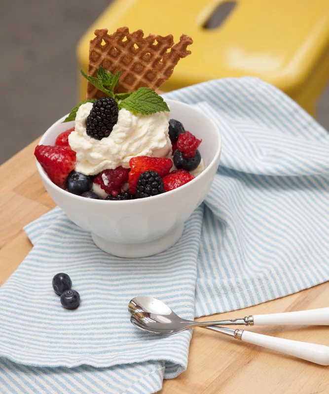 A bowl of mixed berries topped with whipped cream, a sprig of mint, and a waffle piece, placed on a blue-striped cloth on a wooden surface.