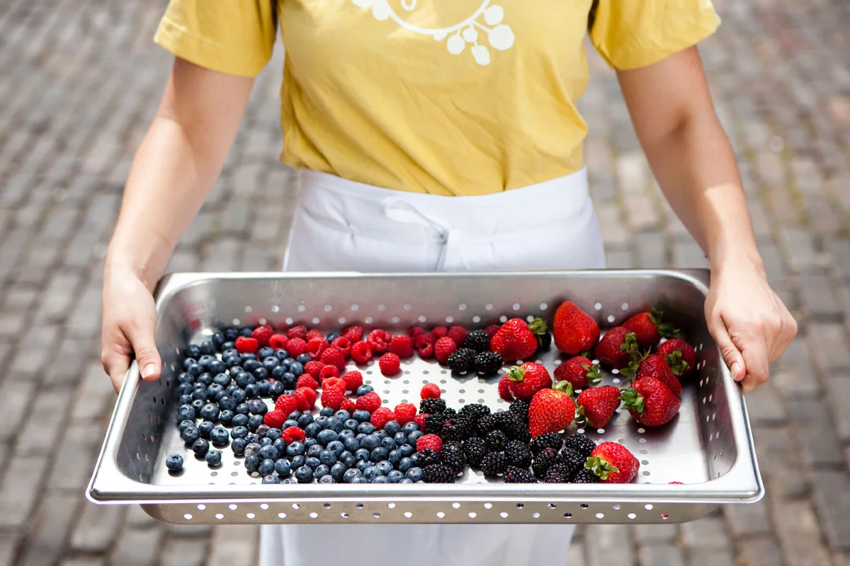 A person holding a metal tray with blueberries, raspberries, blackberries, and strawberries on a cobblestone street.