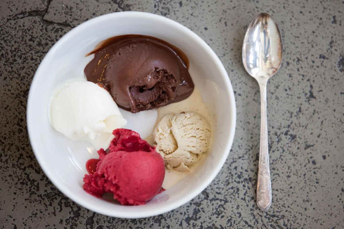 A white bowl of four different scoops of ice cream with a spoon next to it on a speckled gray surface.