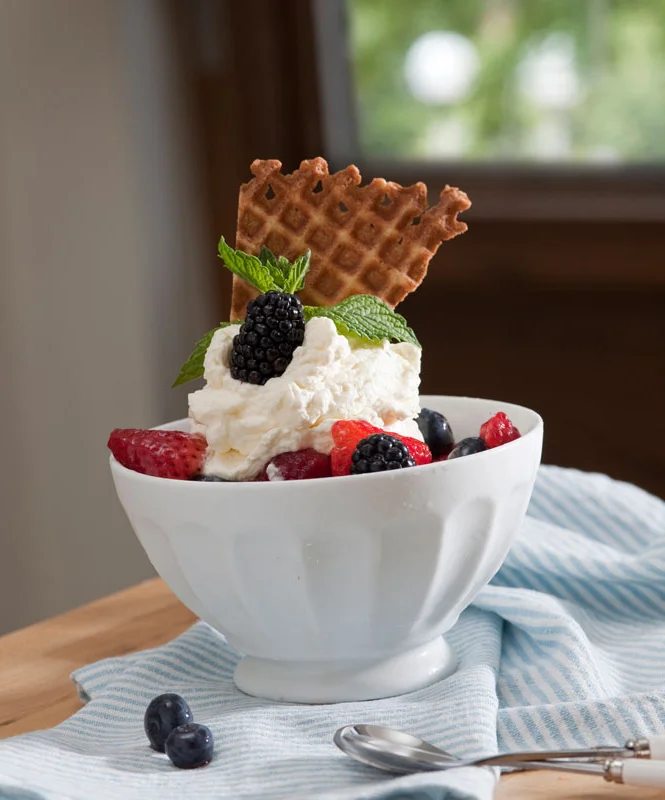 Scoops of vanilla ice cream topped with blackberries, mint leaves, and a waffle cookie, served in a white bowl with assorted berries on a blue and white striped cloth.