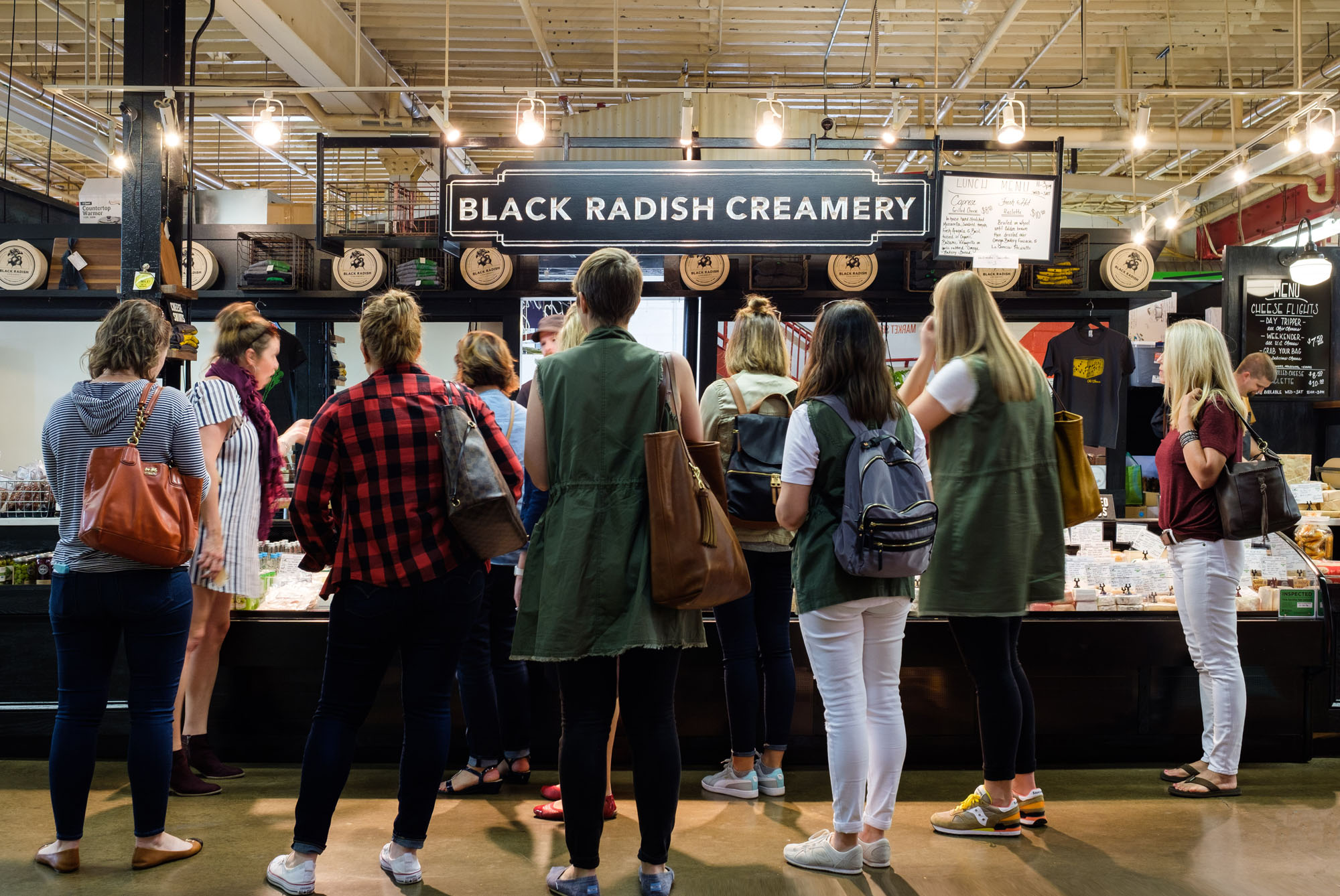People standing in line at a cheese shop named Black Radish Creamery inside a market or grocery store. Customers are looking at products, with various bags on their shoulders.