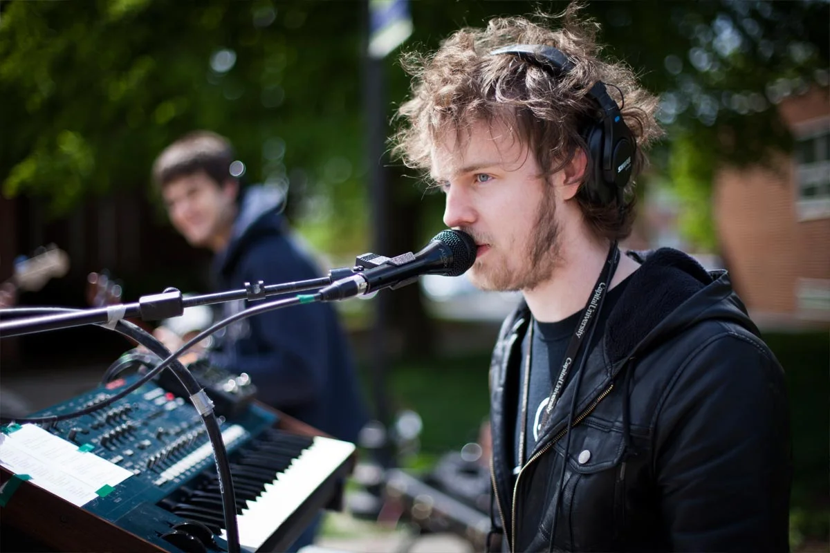 Young man with curly hair and a beard wearing headphones, singing into a microphone while playing a keyboard outdoors, with a blurred young man in the background.