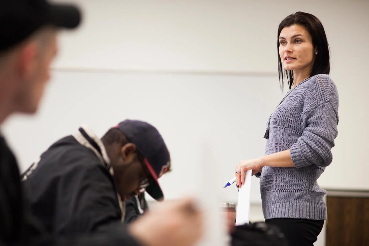 A woman standing at a desk with a whiteboard behind her, holding a dry erase marker and speaking to a group of people sitting in front of her in a classroom or seminar setting.