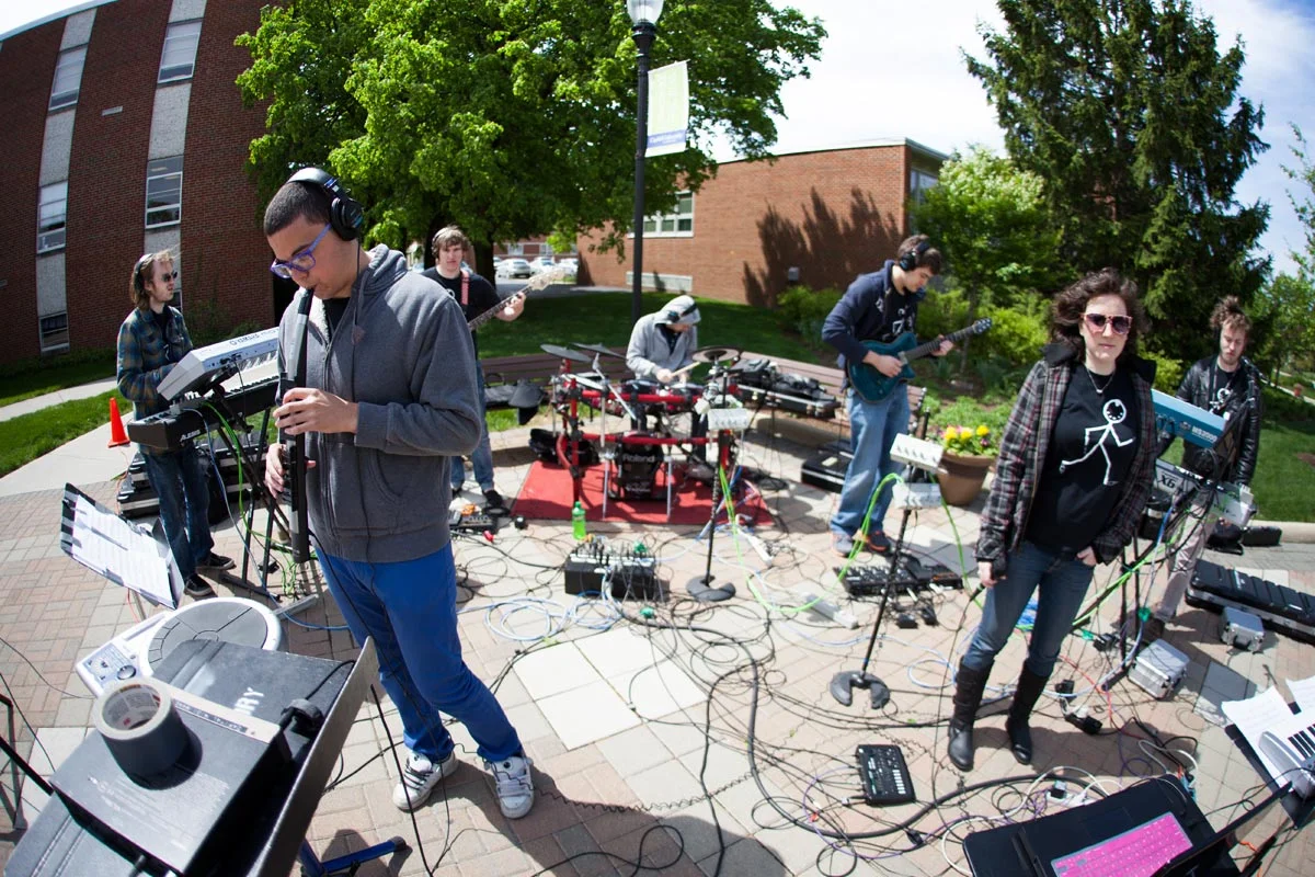 A band of six young people performing outdoors on a brick patio with various musical instruments and equipment. They are surrounded by trees and a building, with greenery in the background.