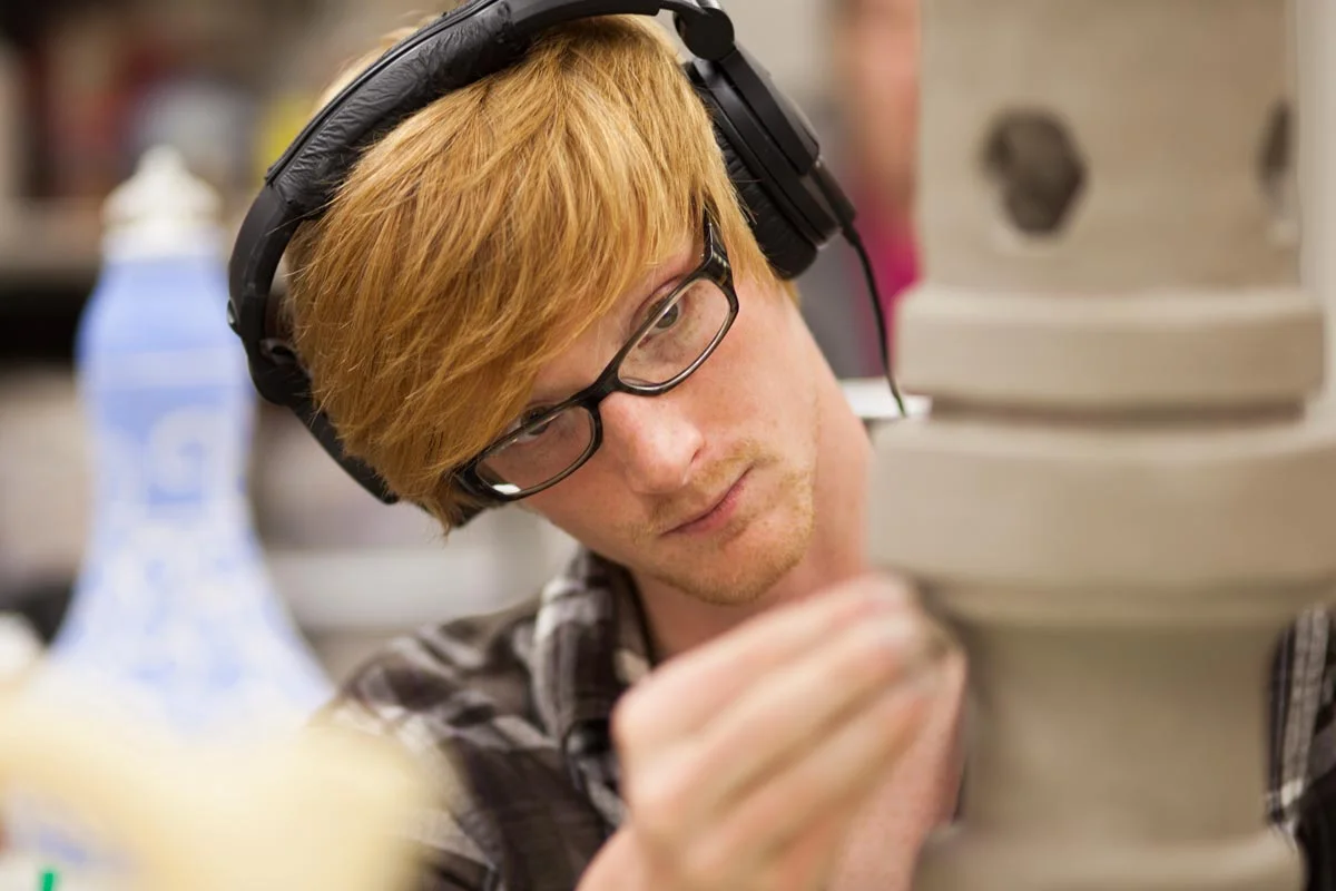 Young man with red hair, glasses, and headphones examines pottery or ceramic item in a workshop or studio.
