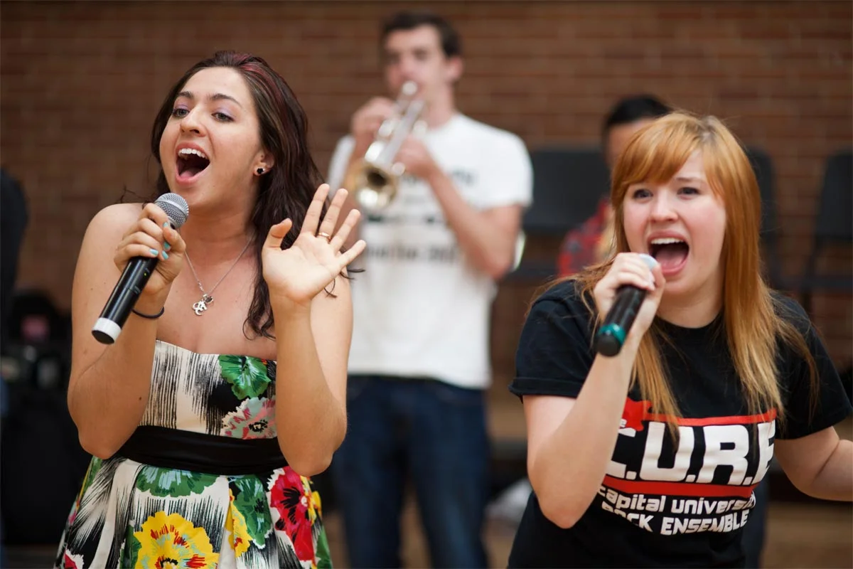 Two women singing into microphones, one with a colorful floral dress and the other with a black T-shirt, while a man in the background plays a trumpet.