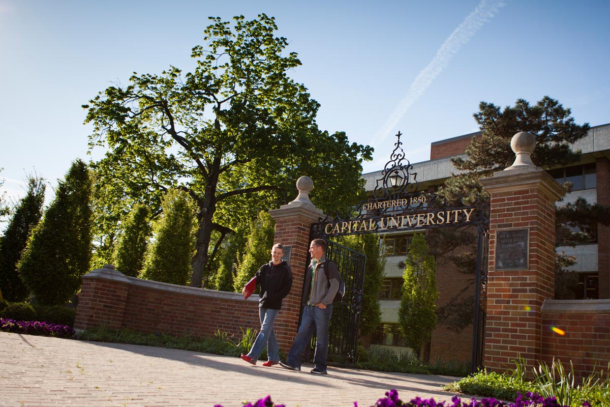 Two students walking out of the gated entrance of Capital University with a large tree and campus building in the background on a sunny day.