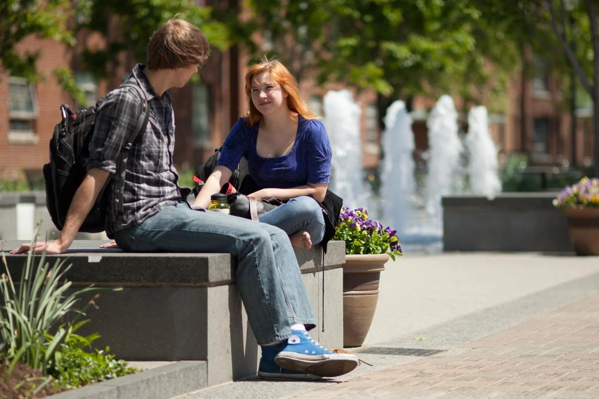 A young man and woman sit and talk on a concrete bench in a park with fountains in the background on a sunny day.