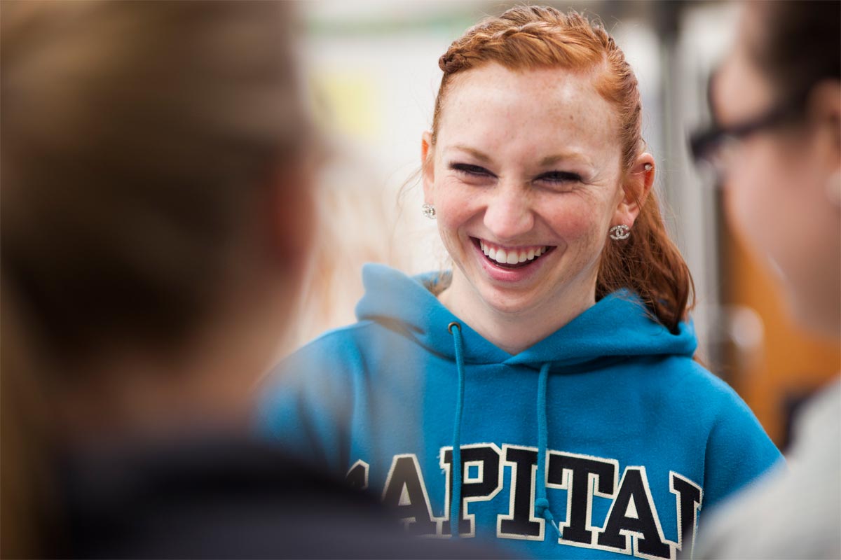 A young woman with red hair smiling and talking to two other people in a social setting, wearing a blue hoodie.