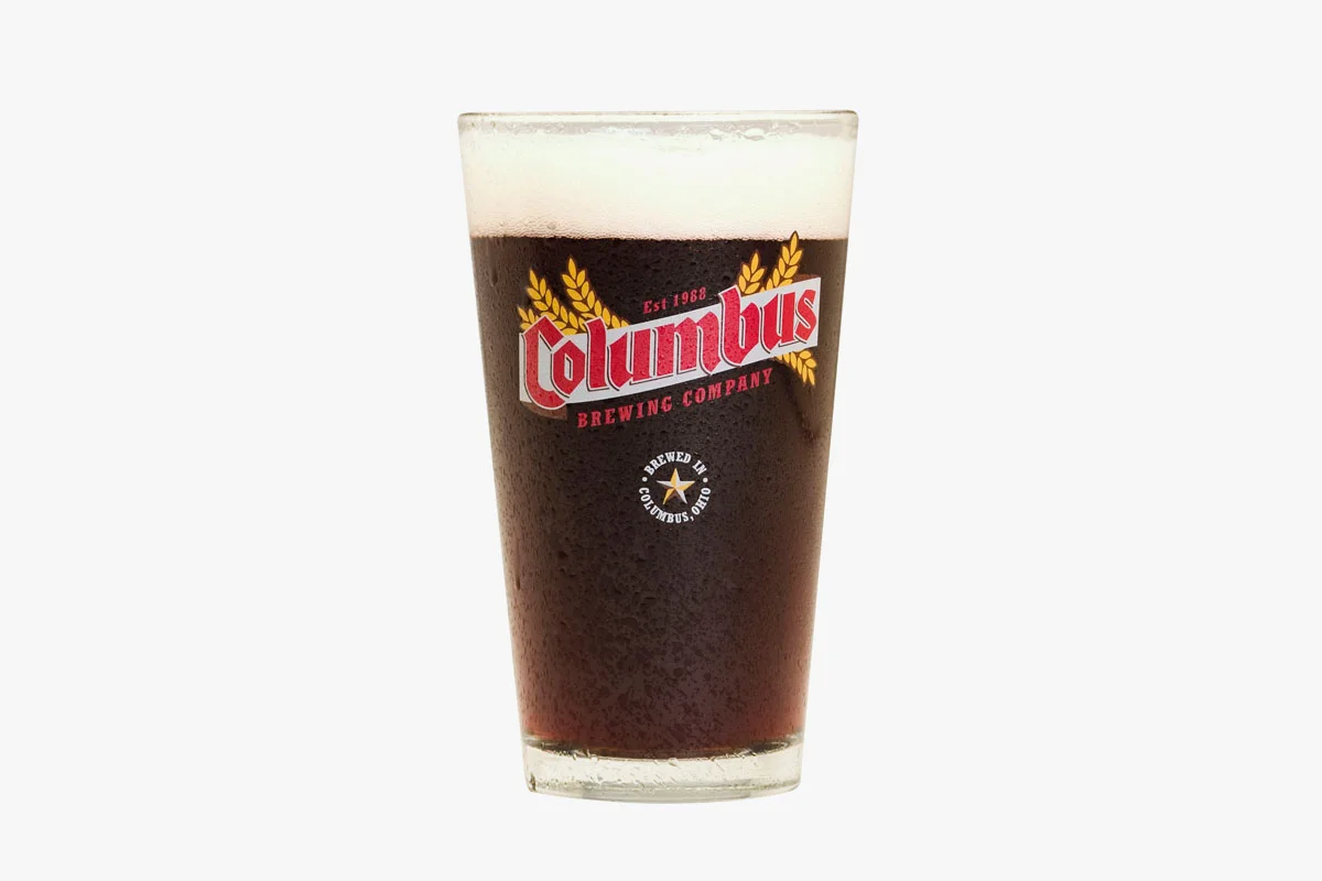 A glass of dark beer with foam head, branded with Columbus Brewing Company logo and text, on a white background.