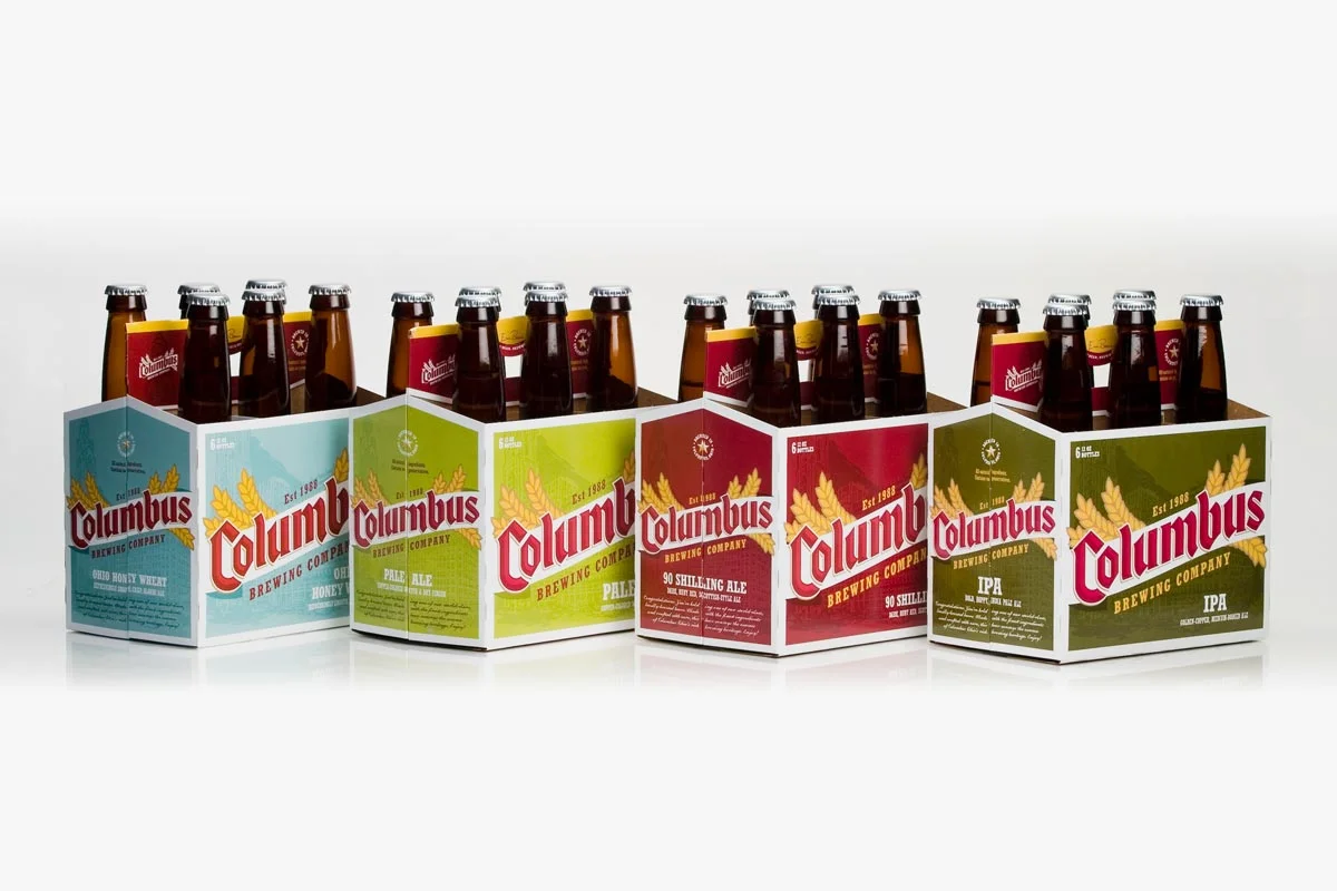 Six packs of Columbus Brewing Company beer bottles with different labels, including Ohio Honey Wheat, Pale Ale, 90 Shilling Ale, IPA, and an unknown label, displayed against a white background.