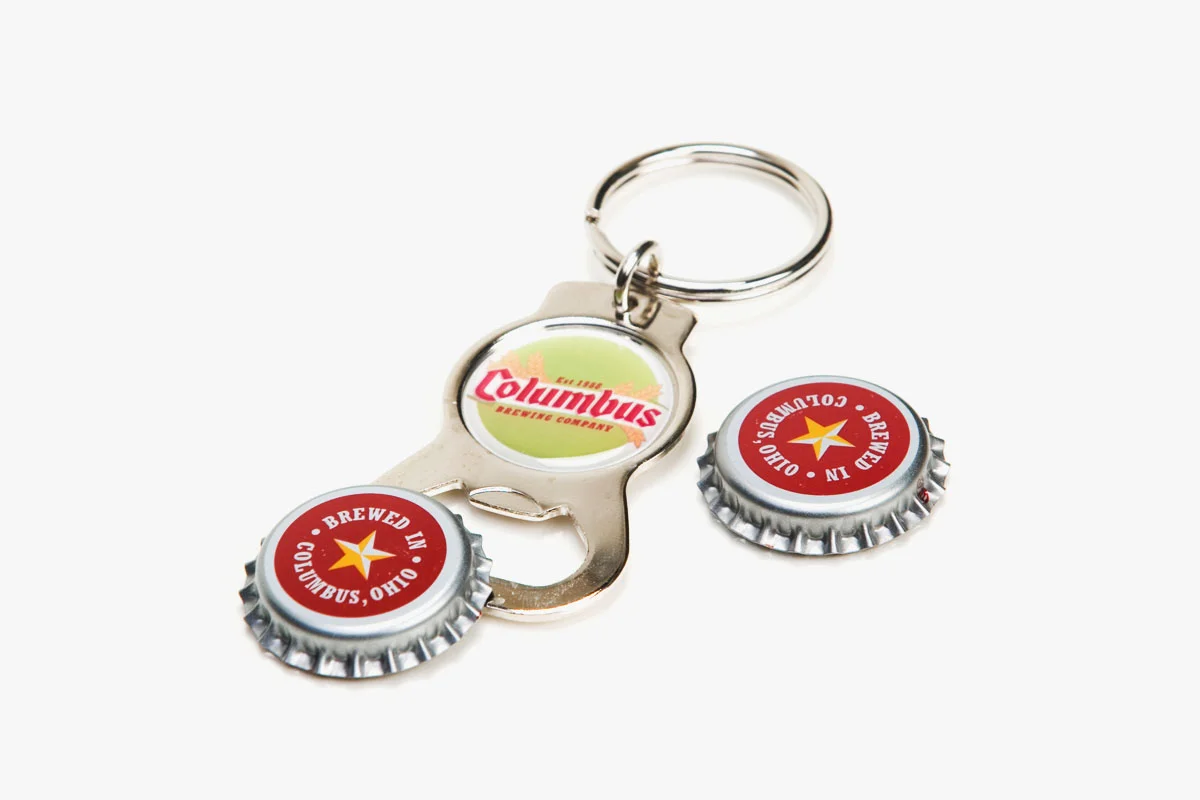 Keychain with two bottle caps featuring Columbus Brewing Company's logo and a metal opener with a logo. The background is white.