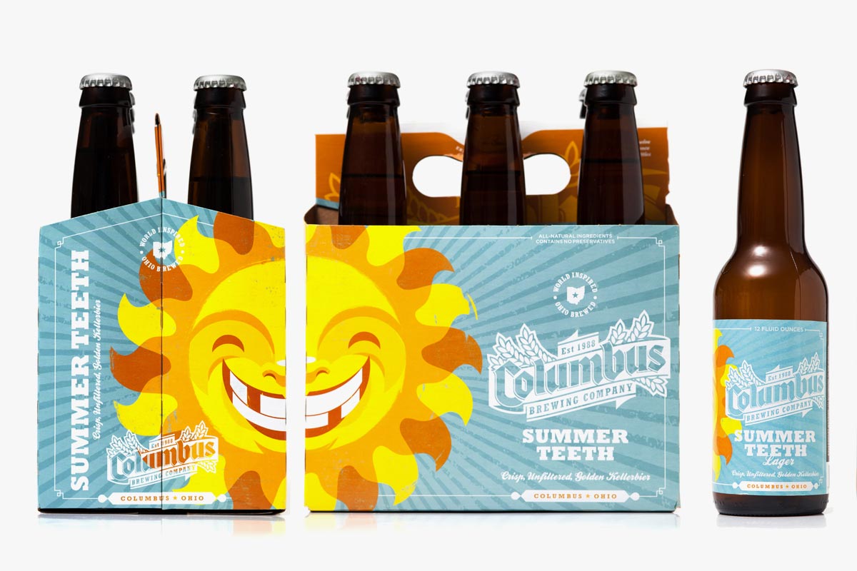 Six bottles of Columbus Brewing Company Summer Teeth beer, with a colorful six-pack carrier decorated with a smiling sun illustration, placed beside a single bottle.