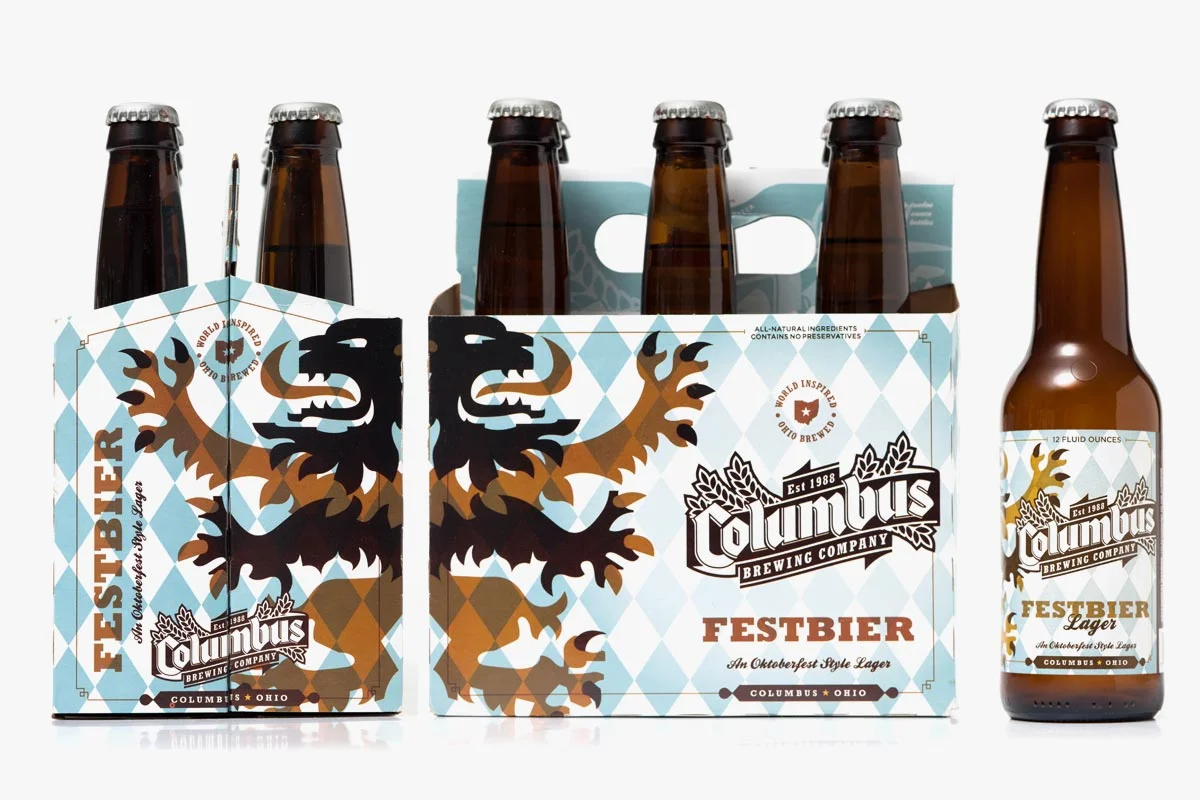 Six bottles of Columbus Brewing Company Festbier beer in a six-pack carrier, with one bottle outside the pack. The packaging and bottle label feature a brown and black eagle with a blue and white diamond pattern background.