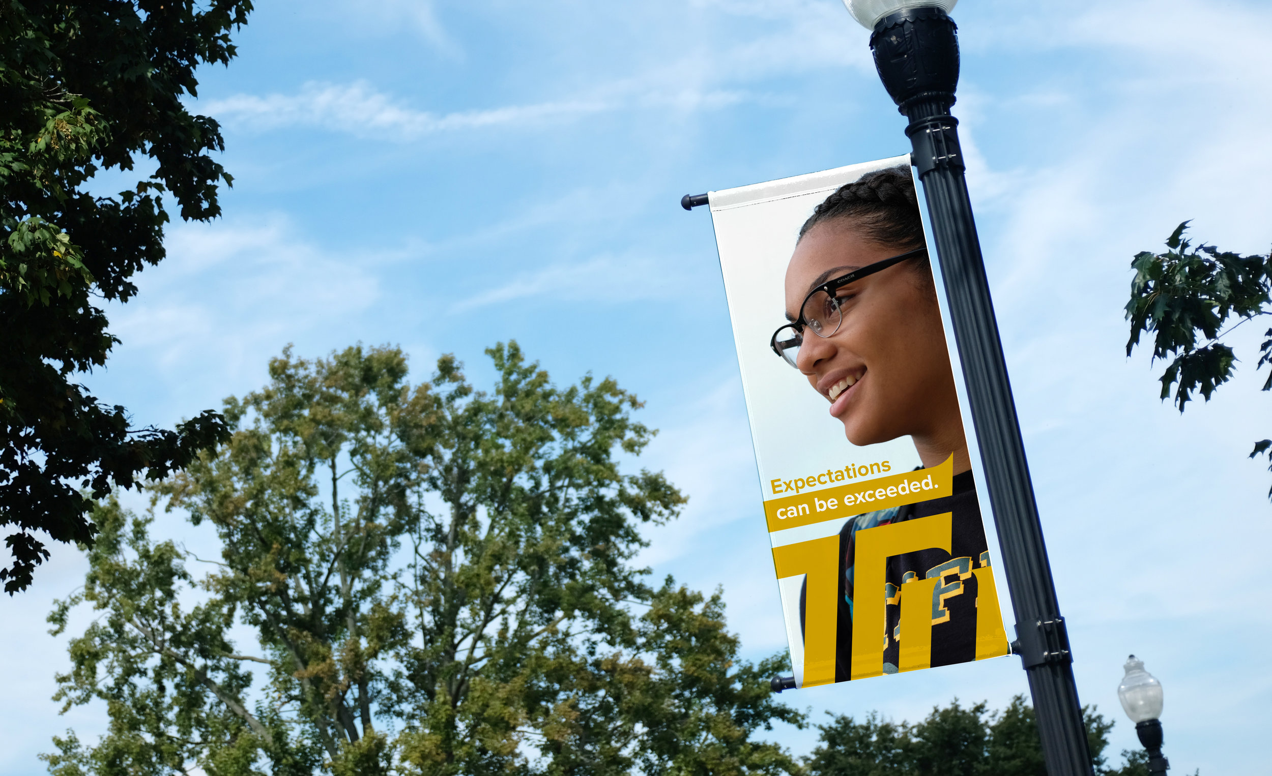 A person with glasses smiling, leaning on a street banner, with trees and blue sky in the background. The banner displays the words 'Expectations can be exceeded' and part of a large design.
