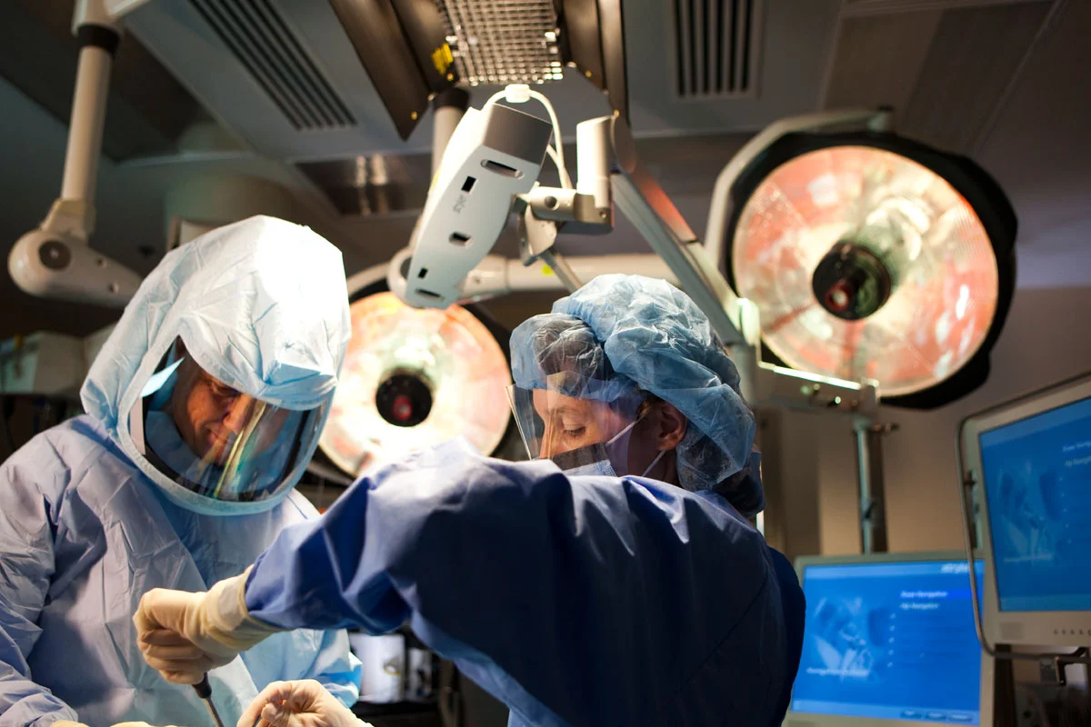 Two surgeons in protective gear performing surgery in an operating room with overhead lights and monitors.