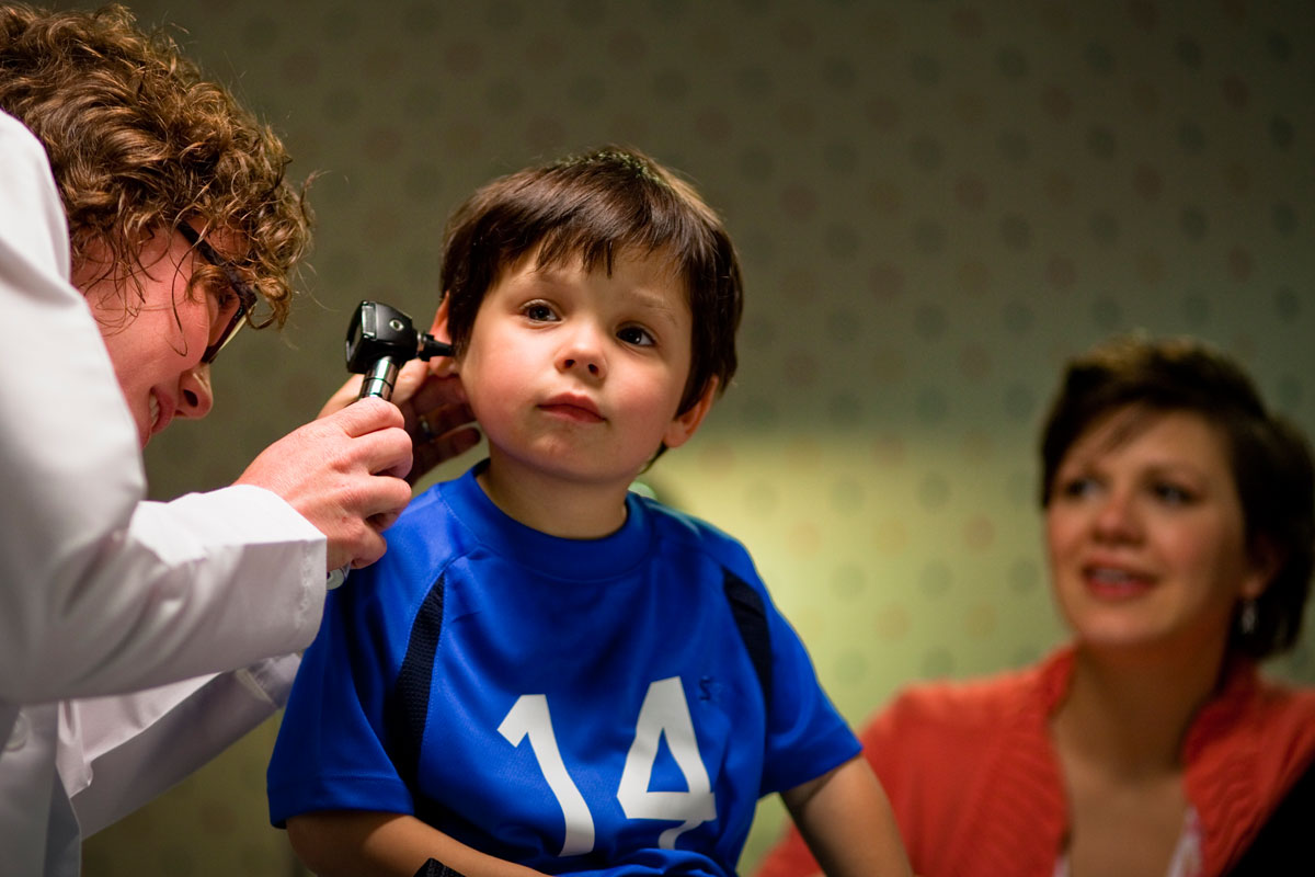 A young boy in a blue sports shirt having his ear examined by a female doctor with a medical instrument, while a woman in an orange sweater watches.