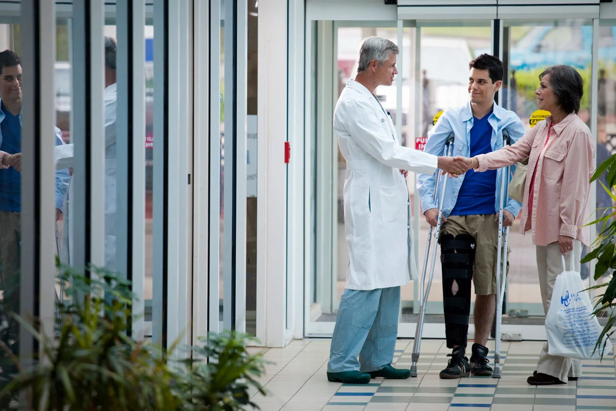 A doctor shaking hands with a woman, who is accompanied by a man with a crutch, at hospital entrance. The man has a leg cast and is wearing a knee brace. Two other medical staff are present nearby.