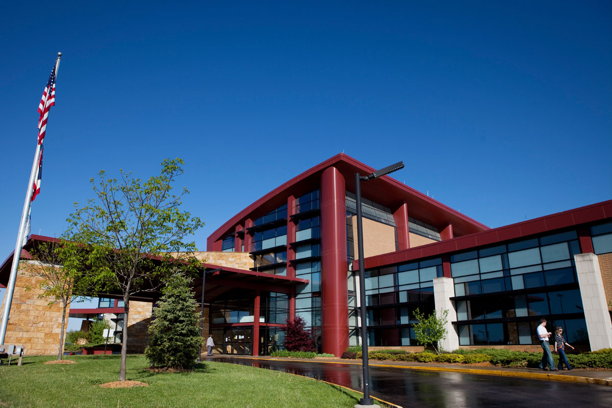A modern multi-story building with red, glass, and brick exterior, surrounded by trees, a flagpole with an American flag, and people walking on a sidewalk against a clear blue sky.