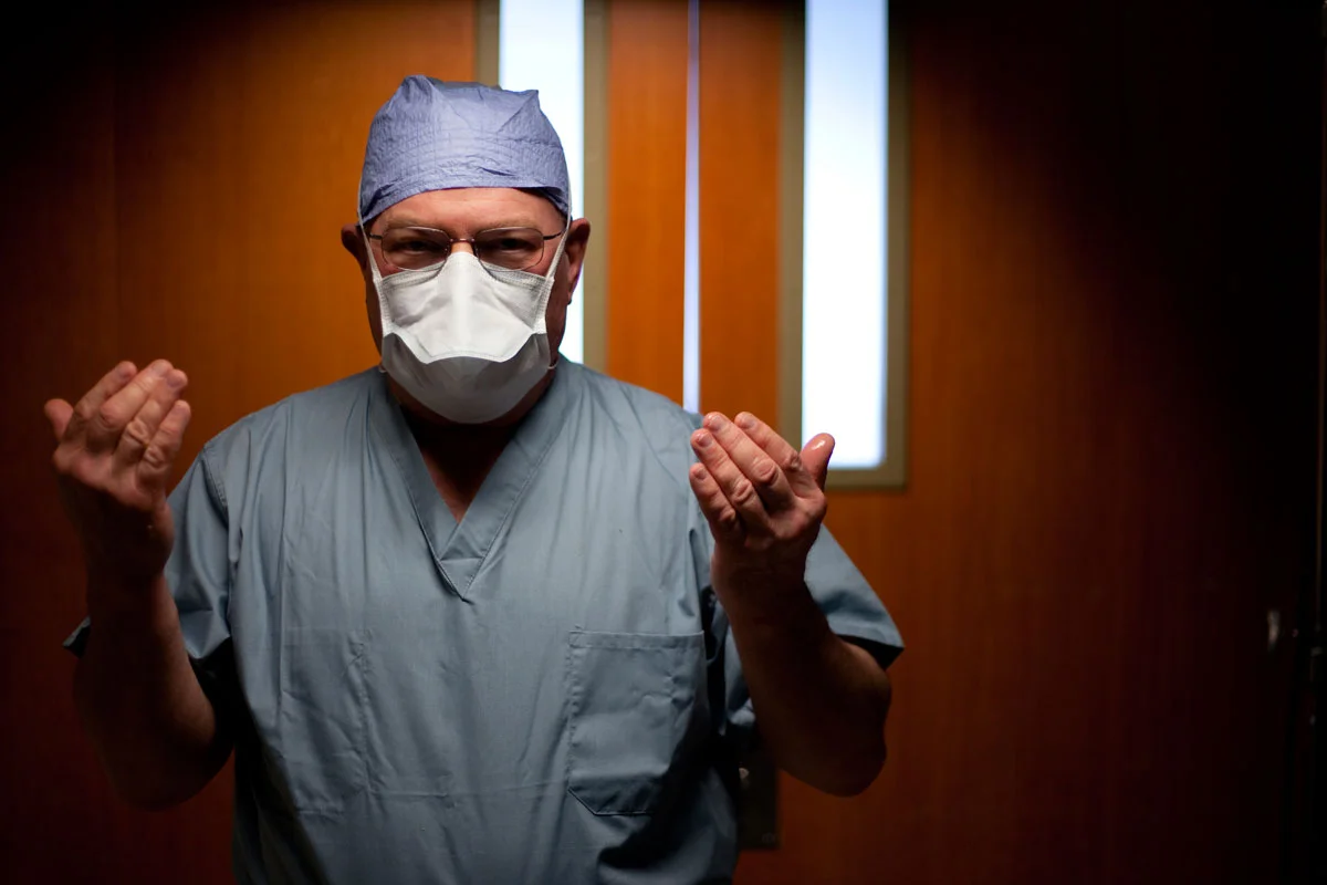 A surgeon in scrubs, mask, and cap standing in a room with wooden walls and a window.