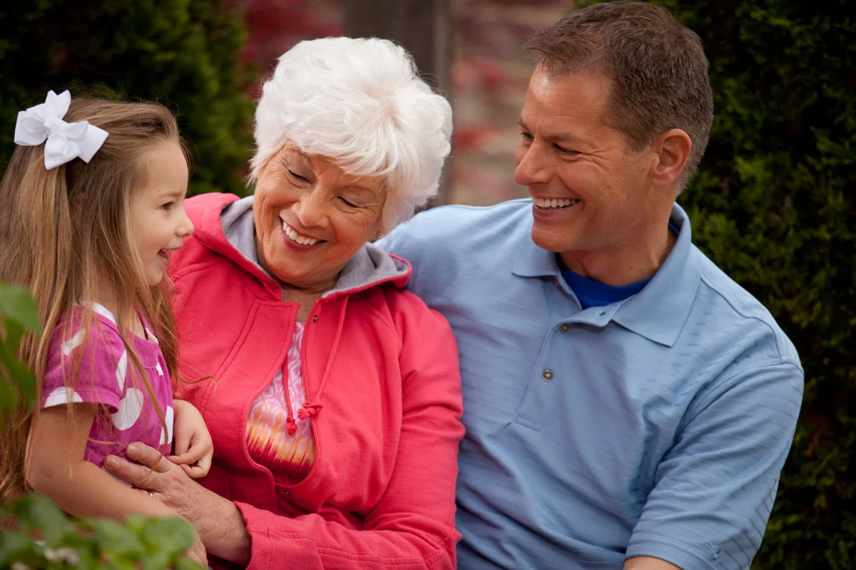Grandparents, a woman with white hair and a man with short brown hair, smiling and sitting outdoors with a young girl with long brown hair and a white bow, all enjoying a happy moment.