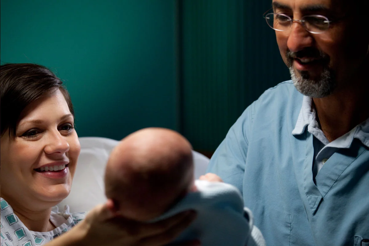A woman in a hospital bed holding a newborn baby, smiling at the baby with a man standing beside her, in a hospital room.
