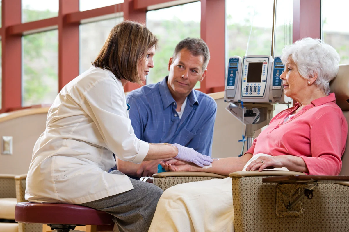A healthcare professional and a man are holding an elderly woman's hand during a medical consultation in a hospital room with large windows.