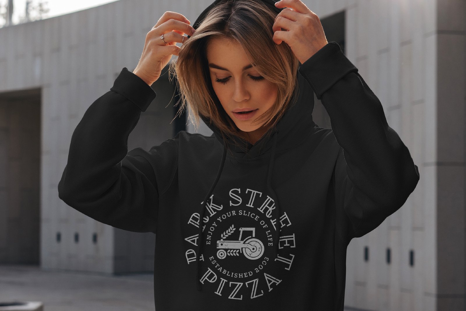 A young woman with shoulder-length hair adjusting her black hoodie in front of a modern building.