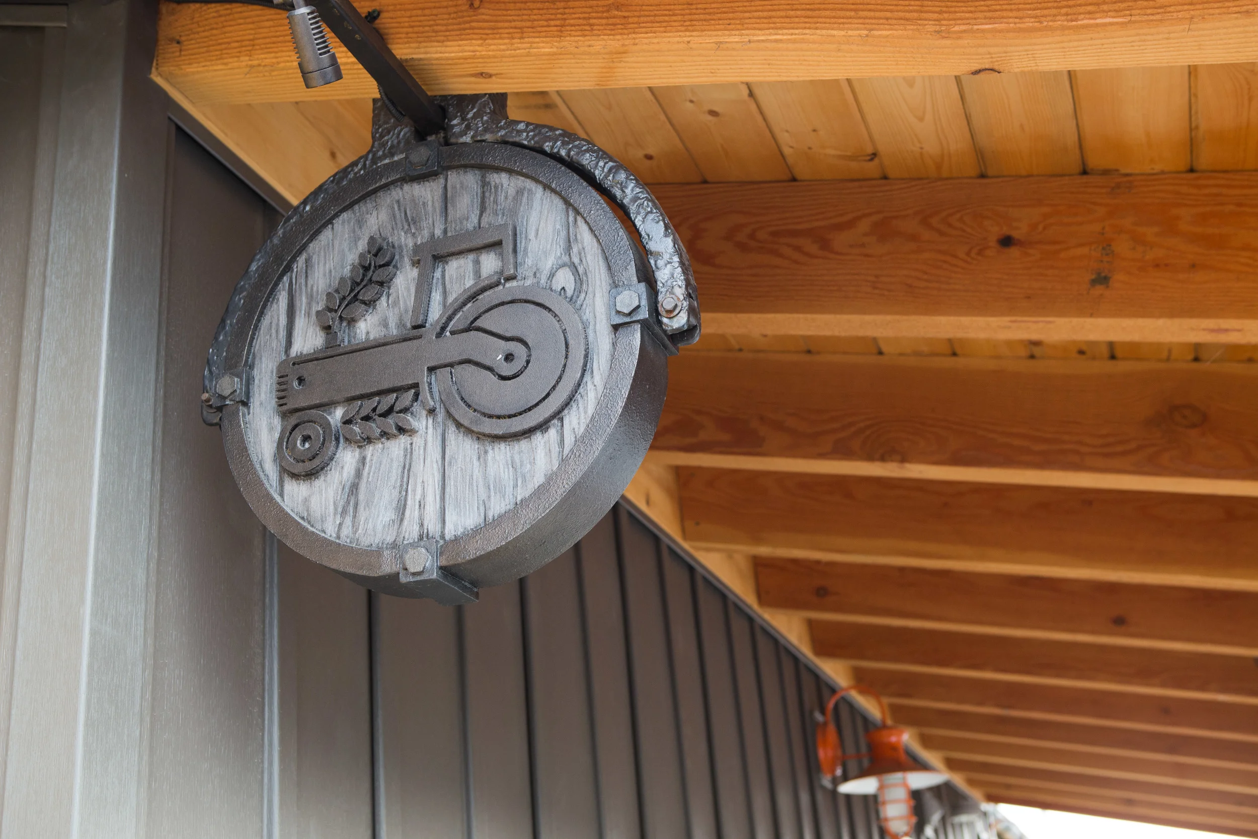 Round decorative wooden sign with metal barn door latch and wheat design, mounted under a wooden roof on a wall.