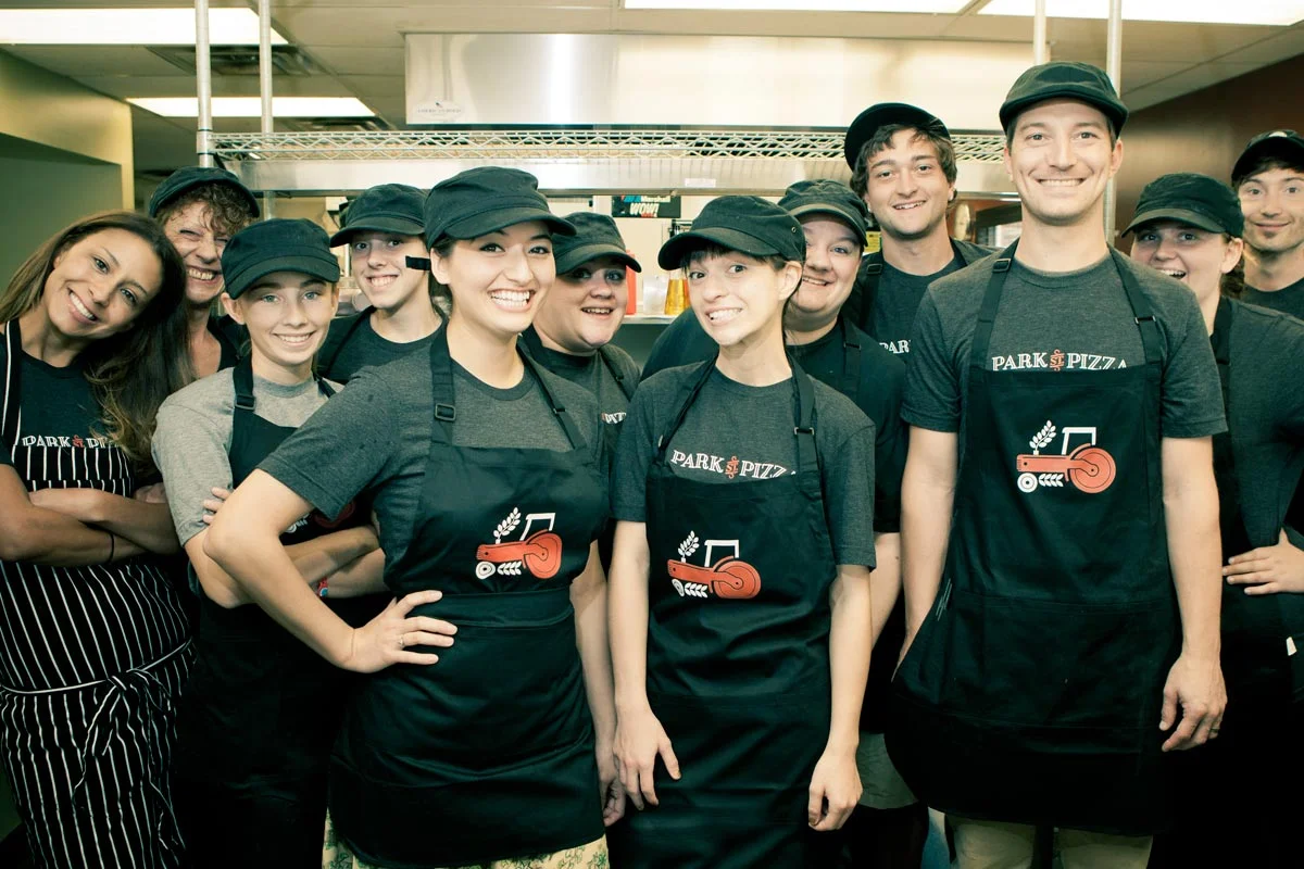 Group of smiling employees wearing matching black aprons and hats in a restaurant kitchen.