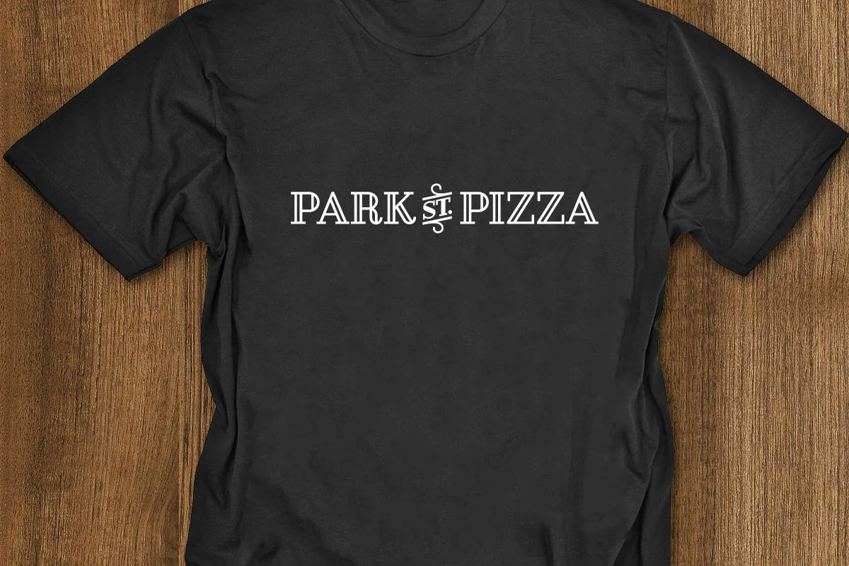 Black T-shirt with white text that reads 'PARK ST. PIZZA' placed on a wooden surface.