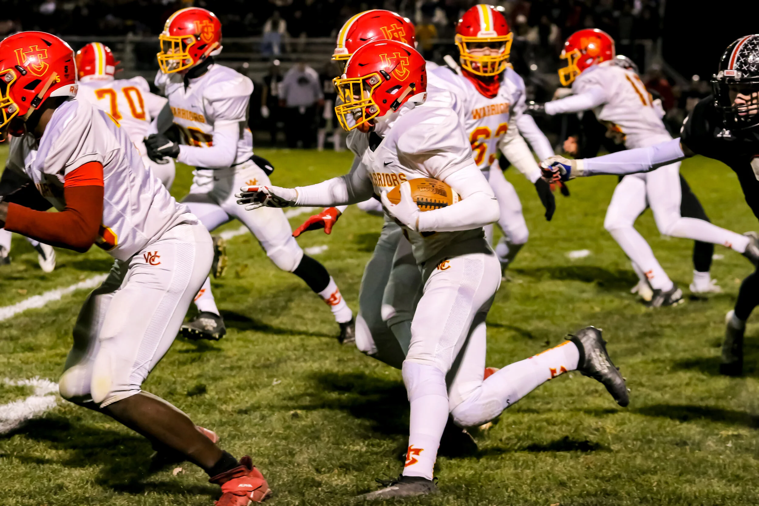 A football player in a white uniform with a red helmet running with the ball during a game on a grassy field at night, surrounded by teammates and opponents.