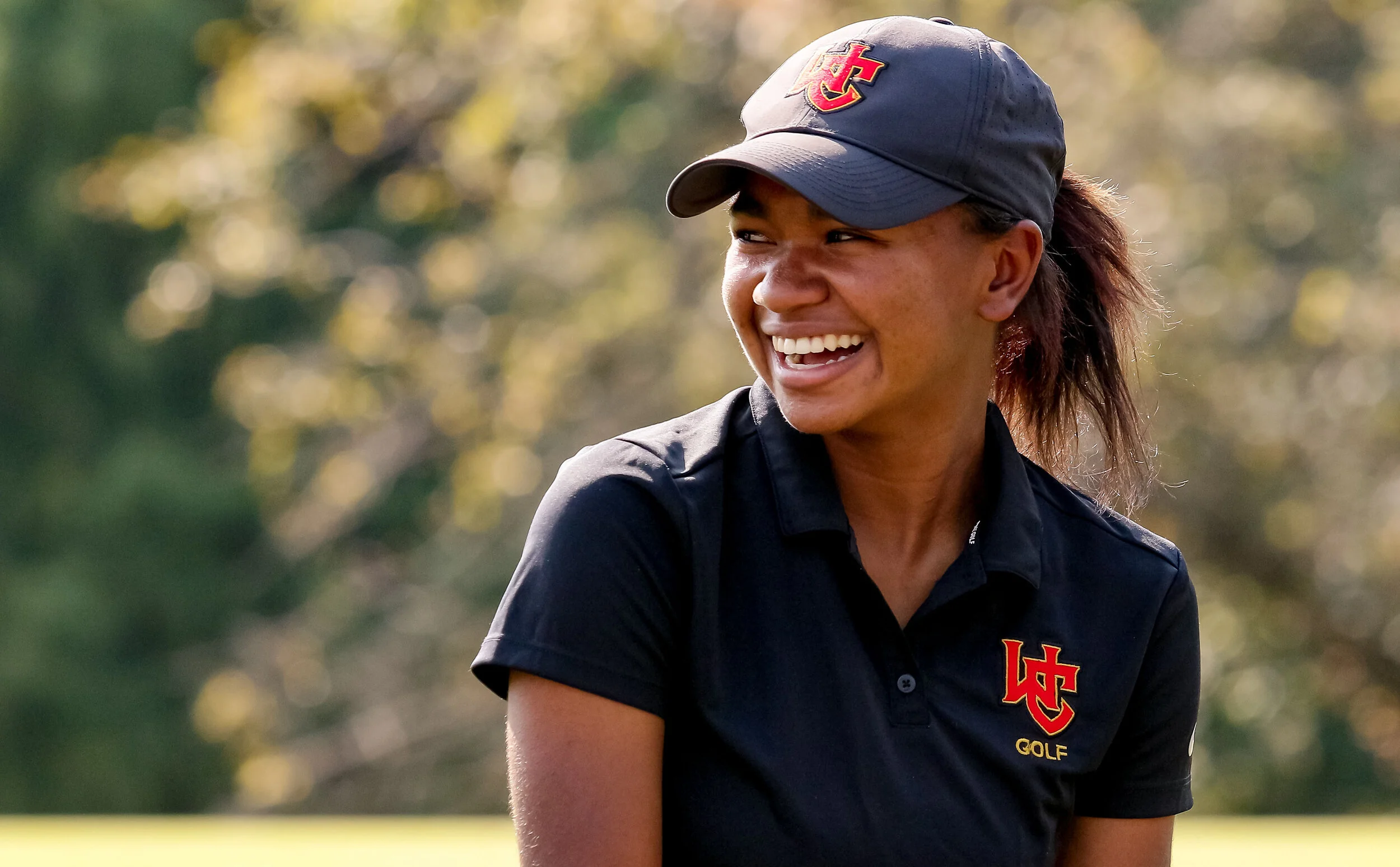 A woman smiling outdoors wearing a navy golf shirt and cap with a red and gold logo.