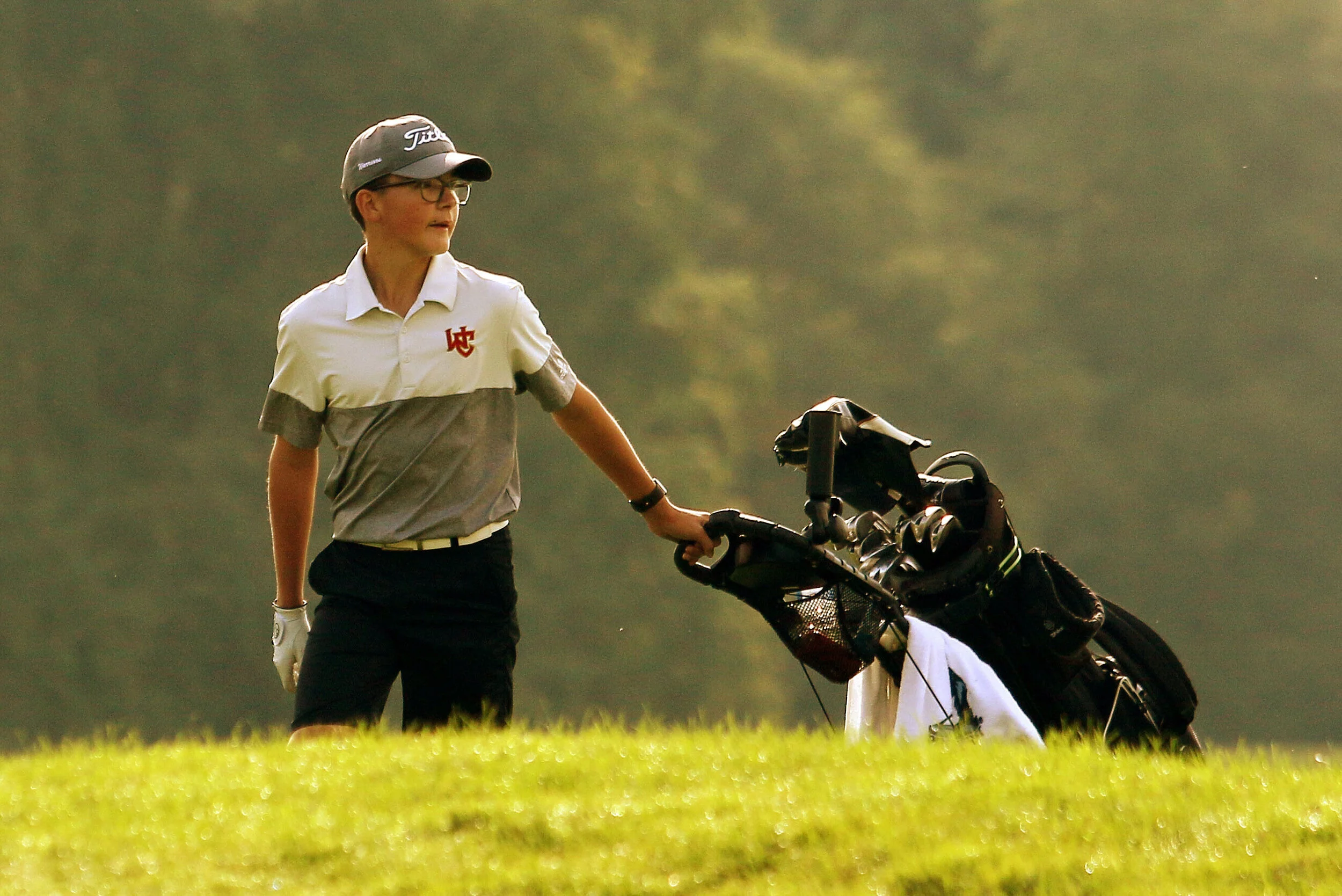 A woman wearing a white and gray golf shirt, black pants, a golf cap, and glasses standing on a golf course holding a golf cart with clubs.