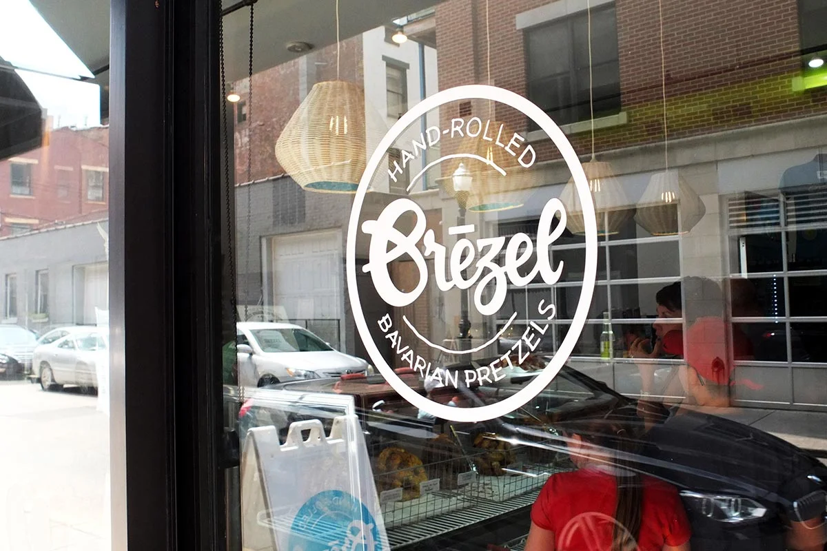 Storefront window with a logo for Gräzel, a Bavarian pretzel shop, featuring a circular design with text and hanging woven lamps inside.