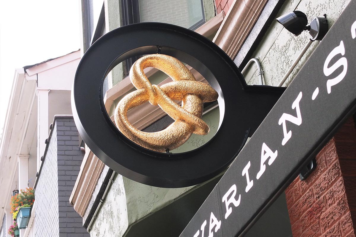 A golden pretzel-shaped sculpture displayed on a black circular stand outside a store with a sign that reads "GOURMET-".