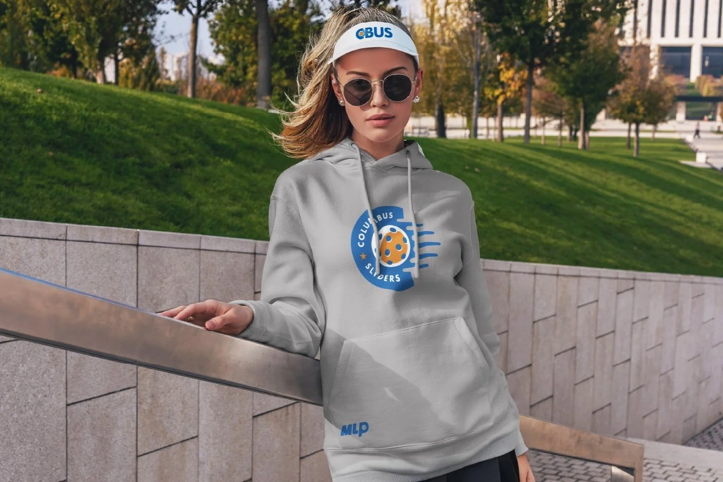 Young woman with sunglasses and a white visor standing outdoors, wearing a gray hoodie with a logo for Columbus Sledders and MLP, in a park with green grass and trees.